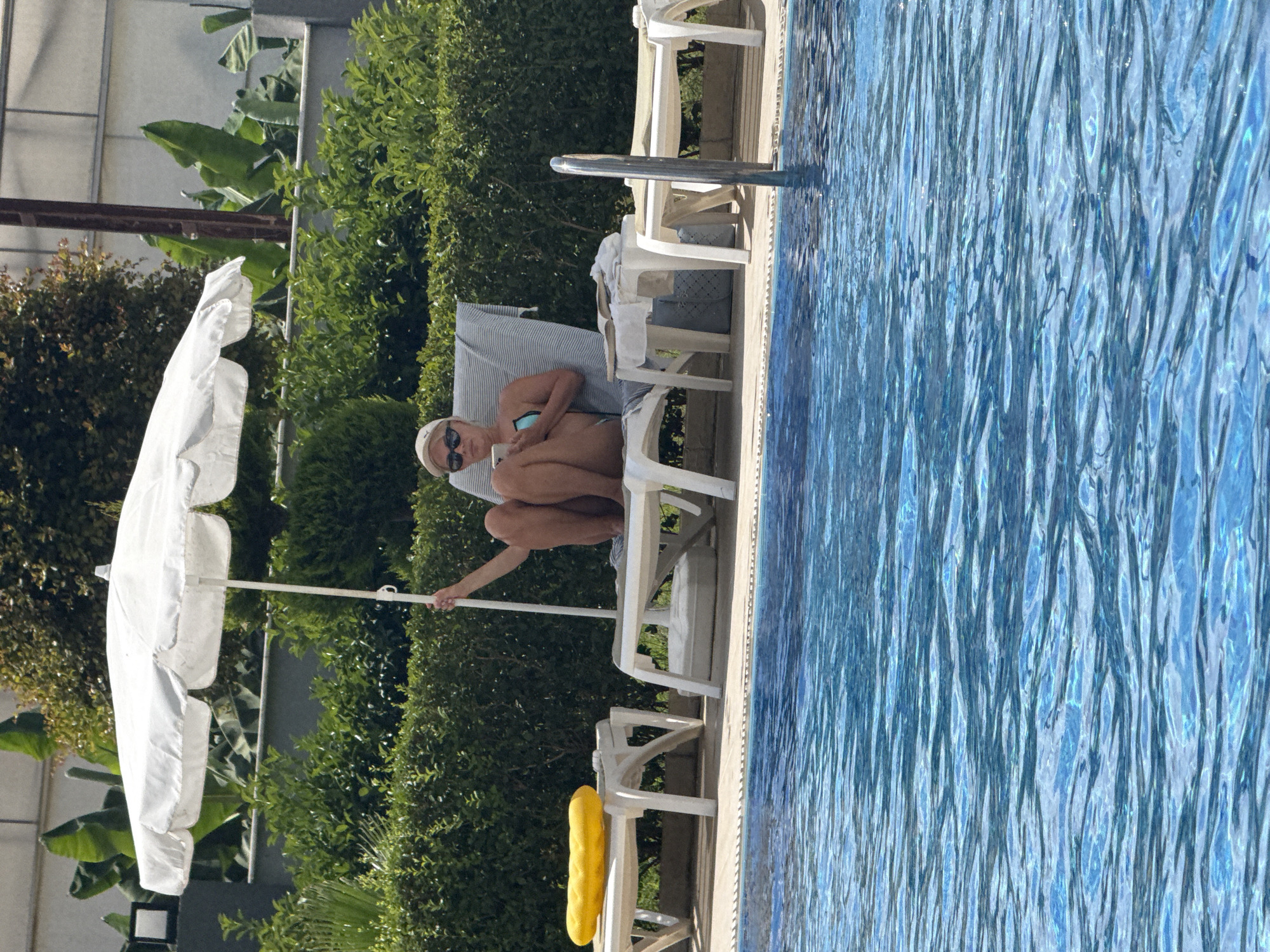 A woman wearing a white cap and sunglasses sits on a lounge chair by a swimming pool. She is wearing a blue bikini and holding a smartphone. To her left, a white umbrella is open. The scene appears to be a sunny day, likely in the afternoon, given the bright sunlight. Lush green bushes are visible behind the lounge chairs. The pool water is a vibrant blue. The overall atmosphere is one of relaxation and leisure, suggesting a holiday or vacation setting. The location context provided is Alanya, Turkey.