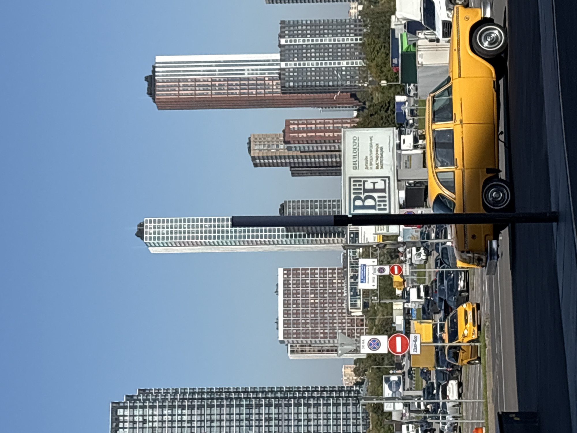An eye-level, medium shot captures a sunny day in Khimki, Russia, with a clear blue sky dominating the background. The scene is an urban landscape characterized by modern high-rise buildings and a busy street. In the foreground, a yellow vintage taxi, likely a GAZ-13 "Chaika," is parked on the right side of the frame, its chrome details and white-walled tires gleaming. To its left, another yellow taxi, a modern minivan, is visible further back in traffic.
The street is filled with various vehicles, including cars and trucks, suggesting a busy traffic environment. Several traffic signs are visible, including a prohibition sign and directional signs, indicating the area is a controlled zone. In the midground, a billboard with Cyrillic text and a logo, possibly advertising a construction or exhibition company, stands prominently.
The background is dominated by a cluster of modern skyscrapers, showcasing a mix of architectural styles and materials. These buildings vary in height and design, some featuring repetitive window patterns and metallic facades, while others display more colorful exteriors. A line of trees separates the road from the buildings, adding a touch of greenery to the urban setting. The overall impression is of a bustling, modern city on a clear, bright day.