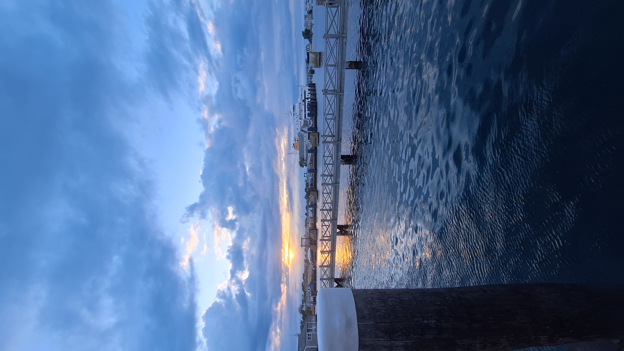 A vertical, eye-level, outdoor shot captures a harbor scene on Nantucket Island, Massachusetts, USA, at dusk. In the foreground, a weathered wooden post with a white cap anchors the left side of the frame. The water, dark blue and rippling, reflects the muted colors of the sky and the setting sun. A wooden pier with a white railing stretches across the middle ground, leading towards a large white ferry docked in the distance. The ferry, with a yellow superstructure, is positioned centrally against a backdrop of scattered clouds tinged with the warm hues of sunset. Beyond the pier, a few buildings with light-colored roofs are visible, suggesting the presence of a coastal town. The sky is a dramatic mix of dark blue and grey clouds, with patches of pale yellow and pink light breaking through, indicating the sun is low on the horizon. The overall mood is serene and twilight-like.