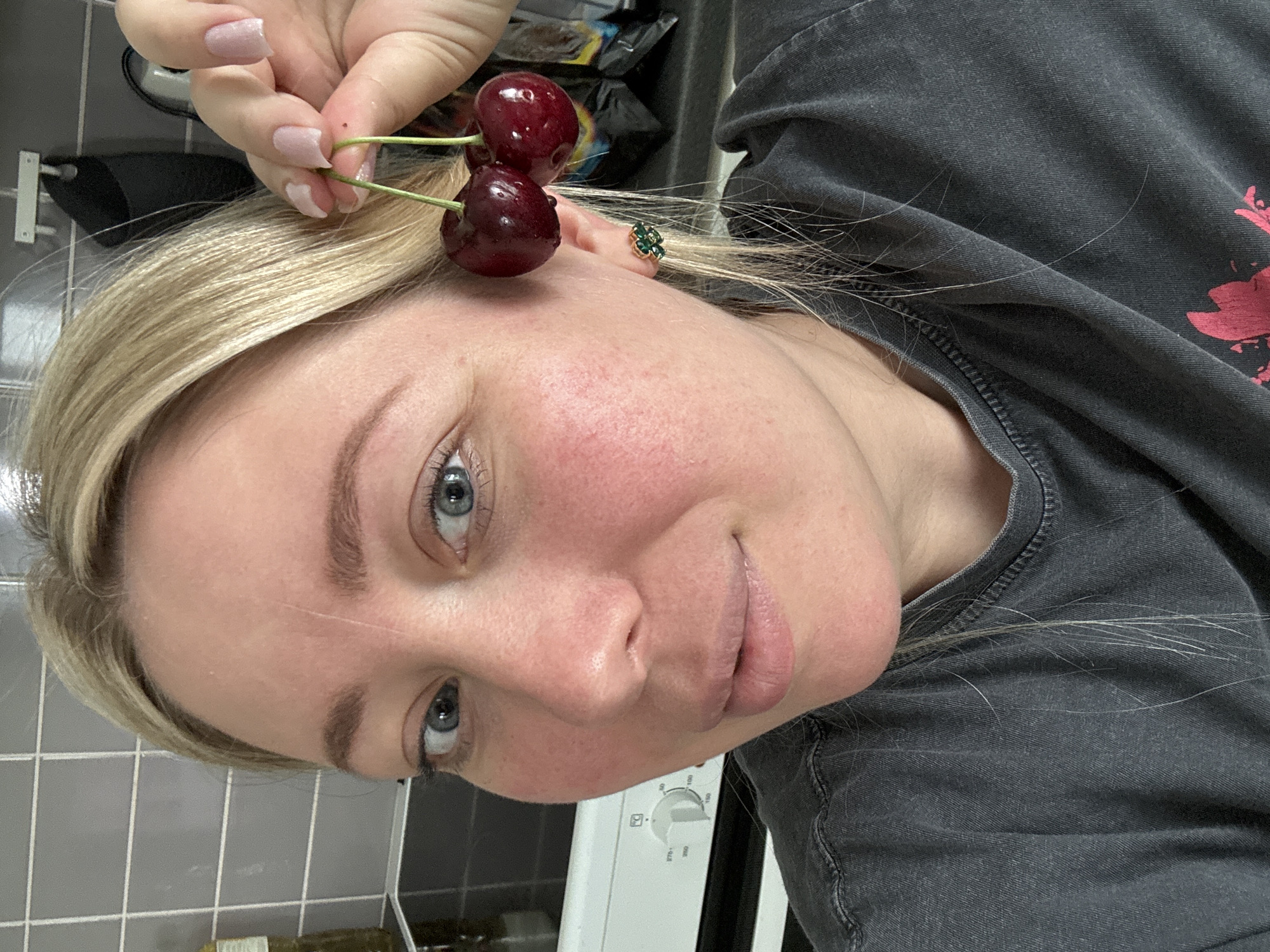 A close-up, head-and-shoulders selfie shows a fair-skinned woman with blonde hair and blue eyes. She is looking at the camera and holding two cherries by their stems near her right ear. She is wearing a dark gray t-shirt and a small green and gold stud earring in her visible ear. The background shows a kitchen with gray tiled backsplash and a white oven.