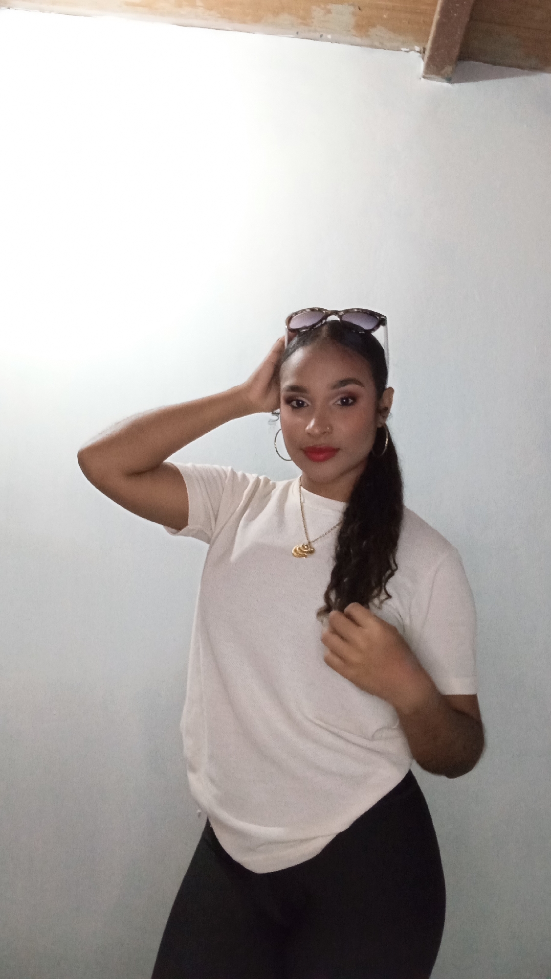A woman stands indoors against a white wall. She is wearing a light-colored t-shirt and black leggings. Sunglasses are perched on top of her head. She has a gold necklace and hoop earrings. Her hair is pulled back into a ponytail. She has one hand on her head and the other resting on her chest. The top of the wall reveals a piece of wooden trim. The image doesn't provide any evidence to suggest that this picture was taken in Juan Griego, Venezuela.