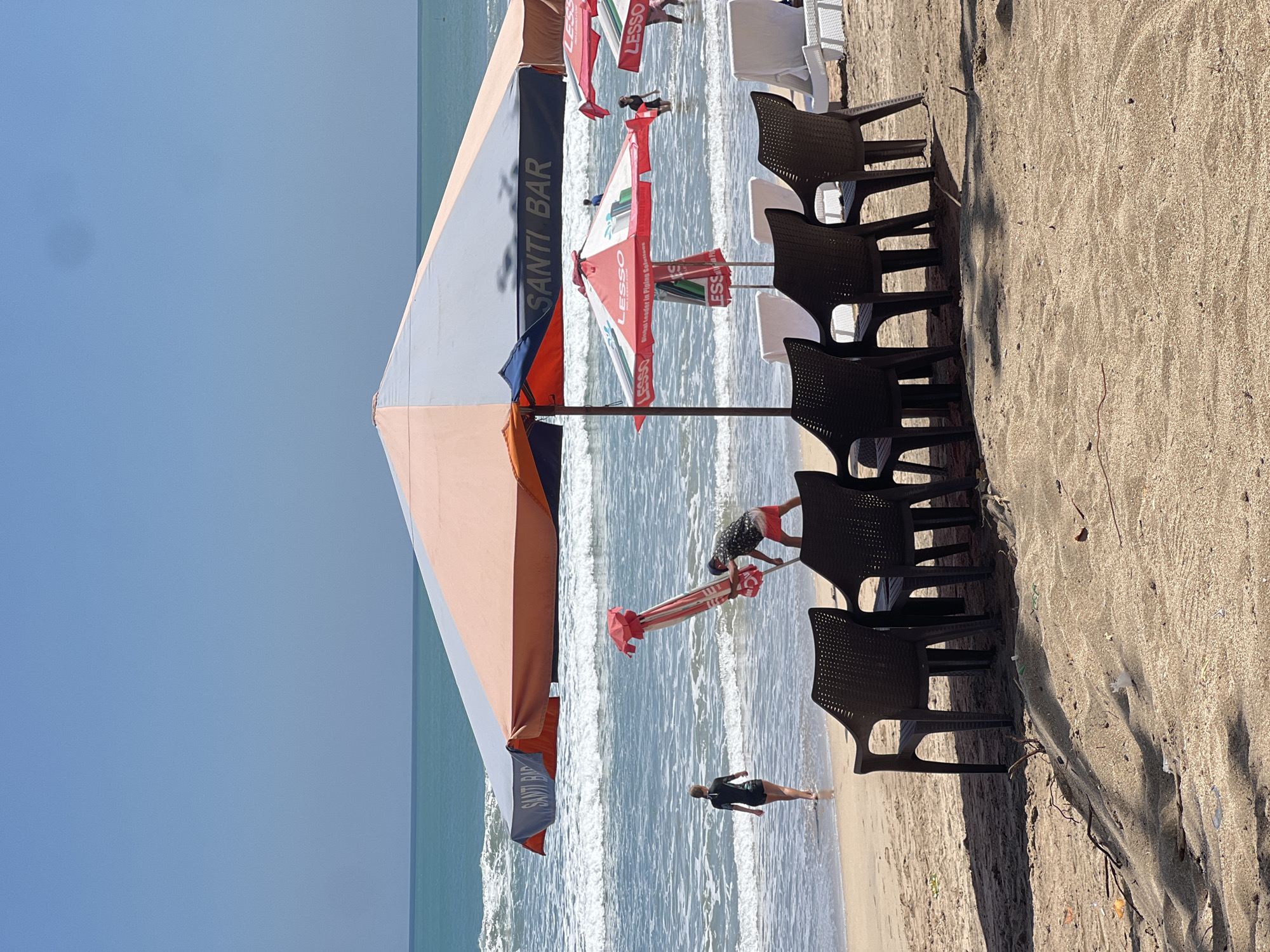 This image depicts a beach scene in Kuta, Indonesia, under a clear blue sky. In the foreground, several dark plastic chairs are arranged on the sandy beach, casting shadows from a large umbrella overhead. The umbrella, predominantly orange and gray, features the text "SANTI BAR" in dark blue lettering.
Further back, the turquoise ocean meets the shore with gentle waves. A person in a black wetsuit walks towards the water. Nearby, a man in red shorts and a patterned shirt is setting up a striped umbrella, holding its pole.
Behind the main umbrella, smaller red umbrellas with "LESSO" branding are visible. Some people are wading in the shallow water. The beach appears to be a public area, likely a tourist spot given the beach chairs and umbrellas. The sand is light brown and shows some debris.