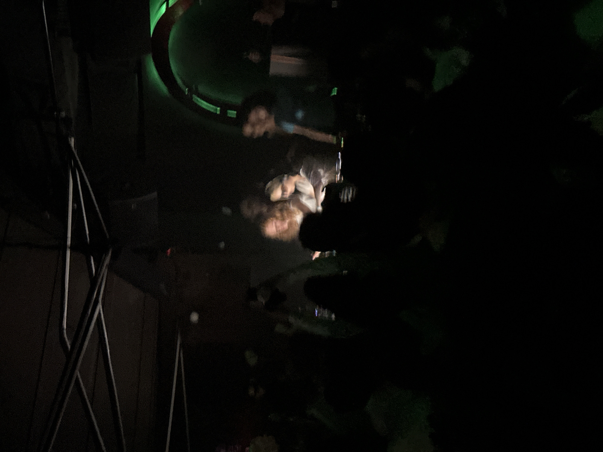 The image depicts an interior scene at the Badaboum nightclub, located at 2 Bis Rue Taillandiers, in the 11th arrondissement of Paris, France. The environment is dark, exhibiting an industrial aesthetic with an exposed ceiling structure composed of metallic trusses and conduits. A crowd of patrons is present, largely visible as silhouettes in the low light, with some individuals holding illuminated mobile phones. In the mid-ground, two women are positioned behind a DJ console, illuminated by direct spotlights. The woman on the left has light-colored hair. The woman on the right wears sunglasses and is actively interacting with the console's controls. To the right of the performers, a prominent archway structure glows with a segmented green neon or LED light. The overall scene captures a live DJ performance in a club setting.