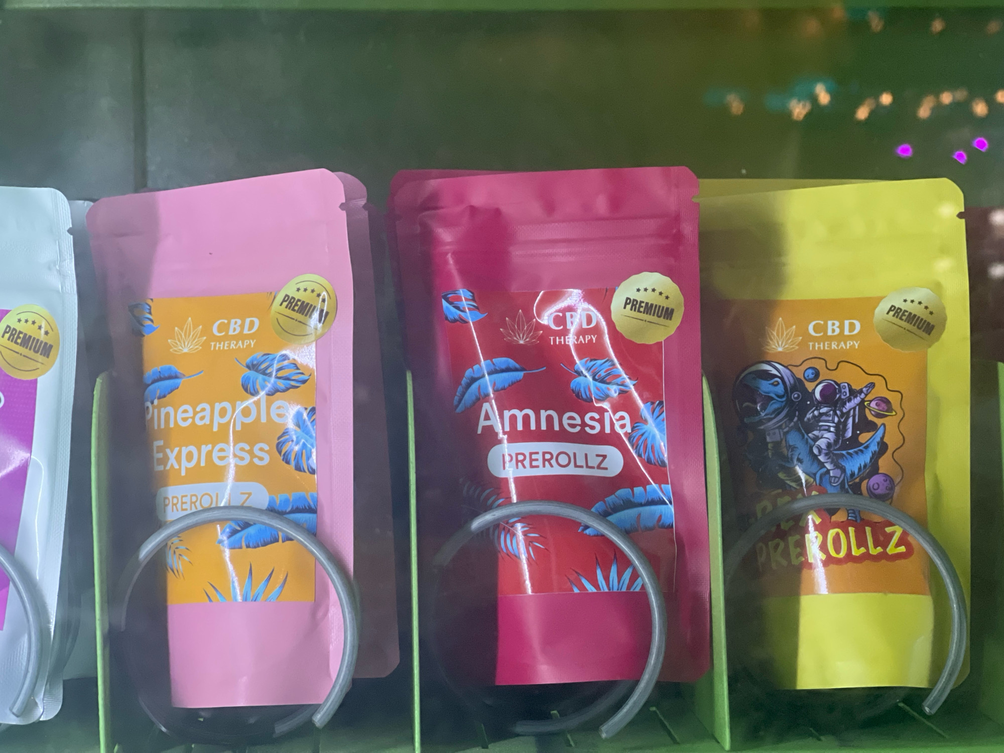 The image shows a close-up of a vending machine stocked with several small, colorful pouches. The pouches are primarily pink, red, and yellow, and appear to be packages of pre-rolled products, likely CBD products, given the "CBD THERAPY" labeling visible on some. Each pouch has various graphic designs, including tropical leaves, a depiction of an astronaut riding a dinosaur, and stars. Circular "PREMIUM" stickers are present on the top right of the pink and yellow pouches. The vending machine has a green exterior and gray metal loops in front of each pouch, typical for dispensing items. The background is blurred and shows green shelving and some out-of-focus lights. The overall impression is that of a retail display for cannabis-related products. The image does not contain any information or visual cues to indicate a specific location within the city of Brno, Czechia.
