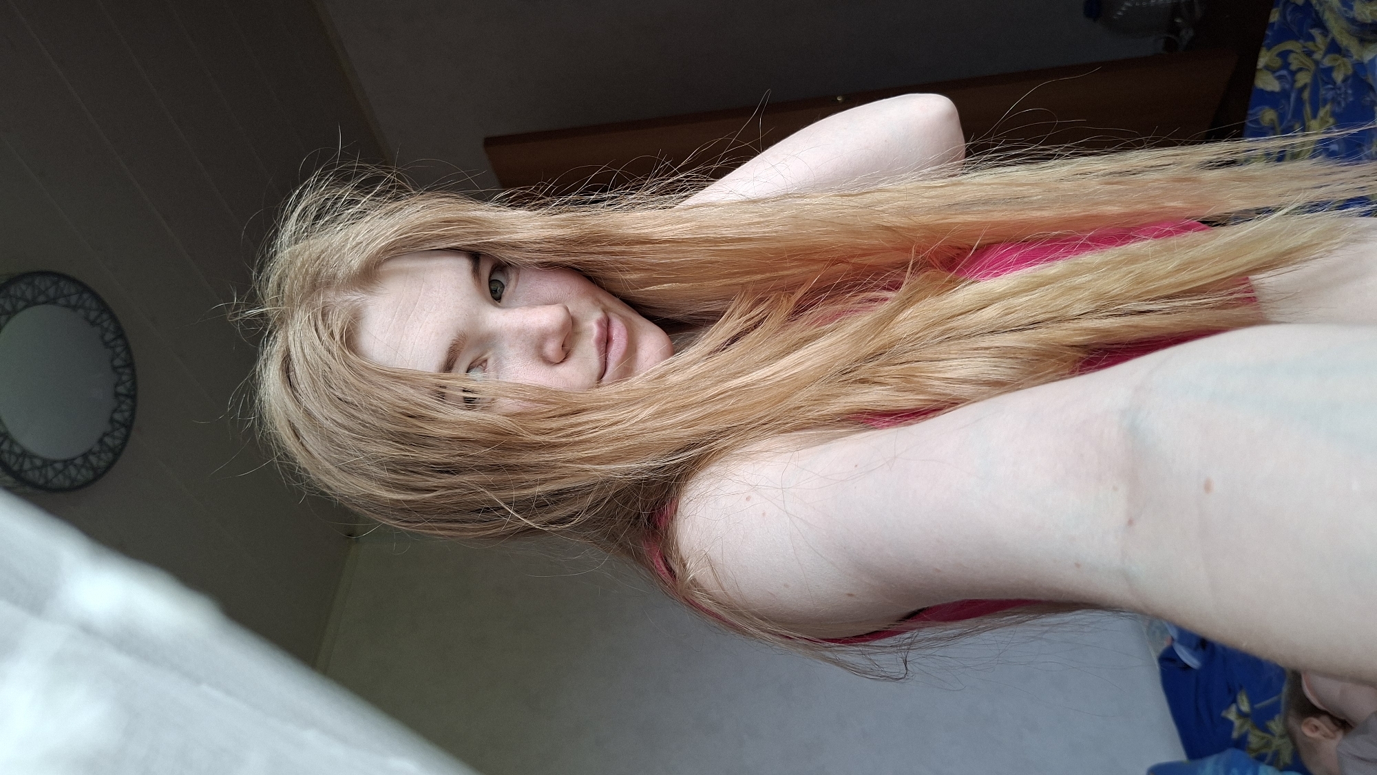 A young woman with long, flowing light blonde or strawberry blonde hair is featured in a close-up, angled indoor shot. She has fair skin, light-colored eyes, and a calm, slightly smiling expression as she looks directly towards the camera. She appears to be lying down, with her voluminous hair fanned out around her head and shoulders, covering parts of her body. She is wearing a bright pink top.
In the dimly lit background, to the upper left, a round mirror with a decorative patterned frame is visible on a wall. A dark wooden headboard or piece of furniture is situated behind her head, and a blue and white patterned fabric, possibly a blanket or curtain, can be seen further in the background to the right. The lighting suggests natural daylight entering the room. No text is visible in the image. The scene takes place indoors, likely a bedroom, in Большой Букор, Russia.