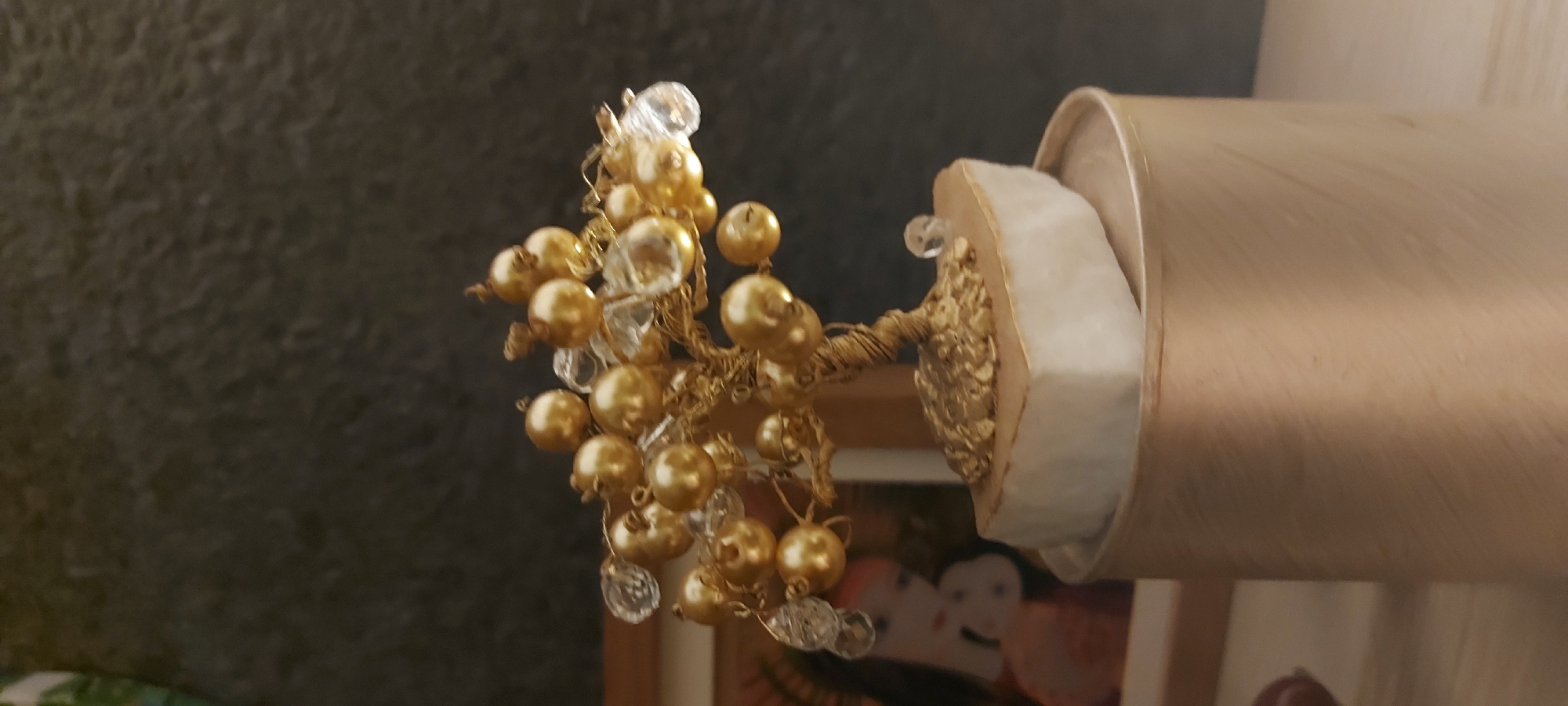 The image shows a decorative golden tree figurine with pearl-like spheres and clear crystals as embellishments. The tree is rooted in a rough, off-white base, which sits atop a cylindrical, pale gold container. In the background, to the left of the tree, a framed picture is partially visible, showing what appears to be a drawing of two figures. The overall setting suggests an indoor display, possibly on a shelf or table, with a dark, textured background. There is no indication of the city of Riga, Latvia, in the image.