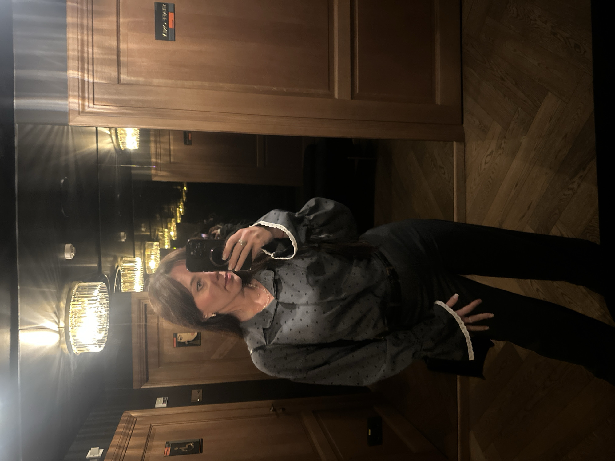 A middle-aged woman stands in a bathroom, taking a selfie with her cell phone. She is wearing a long-sleeved, button-down, black shirt with white trim on the cuffs and collar, and black pants. The bathroom has wooden paneled walls and herringbone wooden floors. There are crystal light fixtures on the wall and various signs on the doors, written in a language other than English, possibly Latvian. The scene suggests a bathroom in a hotel or upscale restaurant in Riga, Latvia. The woman is interacting with her phone, posing for a selfie.