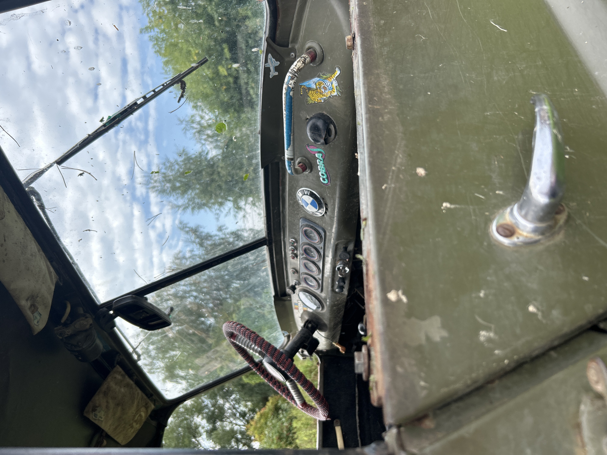 The image shows the interior of a weathered, olive-drab vehicle, likely a military or utility truck, viewed from the passenger side. The dashboard is visible, featuring multiple gauges and a steering wheel covered in a red and black patterned wrap. Several stickers adorn the dashboard, including a BMW logo, a "COBRA" sticker with a snake, and a cartoon tiger holding a flag. A chrome door handle is prominent in the foreground. The windshield is dusty and has a single windshield wiper. Through the windshield, trees and a cloudy sky are visible, suggesting an outdoor setting. The overall condition of the vehicle's interior indicates it is not in active use and has been exposed to the elements. There are no people visible in the image. The location is specified as Kalitsino, Russia, though no specific city landmarks confirm this.