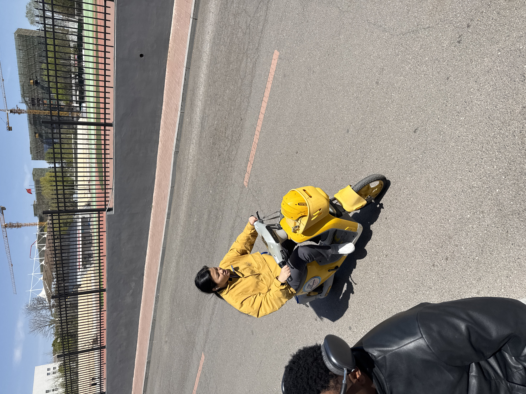 A woman in a yellow jacket rides a yellow scooter on an asphalt road. A construction site, featuring cranes and a building under construction, is visible beyond a black fence. A person wearing headphones and a black leather jacket is partially visible in the lower right corner of the image. A faded orange line is present on the road. The location is Zhongtun, China.