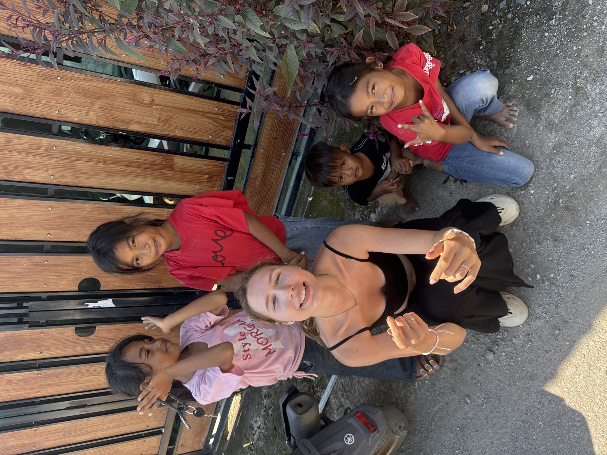 A group of five children and one adult woman are posing for a photo outdoors, in what appears to be Kertasari, Indonesia. The children, four girls and one boy, are positioned around the adult woman. Three girls are standing in the background, two of whom are wearing red t-shirts, and one is wearing a pink t-shirt with the text "Style Morgan" on it. The youngest girl, in the foreground, is wearing a red t-shirt and blue jeans, and she is making a hand gesture. A young boy in a black t-shirt and light brown shorts is crouched next to her. The adult woman, with blonde hair and a black top, is in the center of the frame, and she is smiling and holding her hands up, showing off rings on her fingers. She is wearing a black top and black pants. Behind them is a wooden fence with black metal bars. A portion of a motorcycle is visible on the left side of the frame. The ground is covered in gravel and dirt. A plant with purple leaves is growing on the right side of the frame. The scene is illuminated by natural sunlight.
