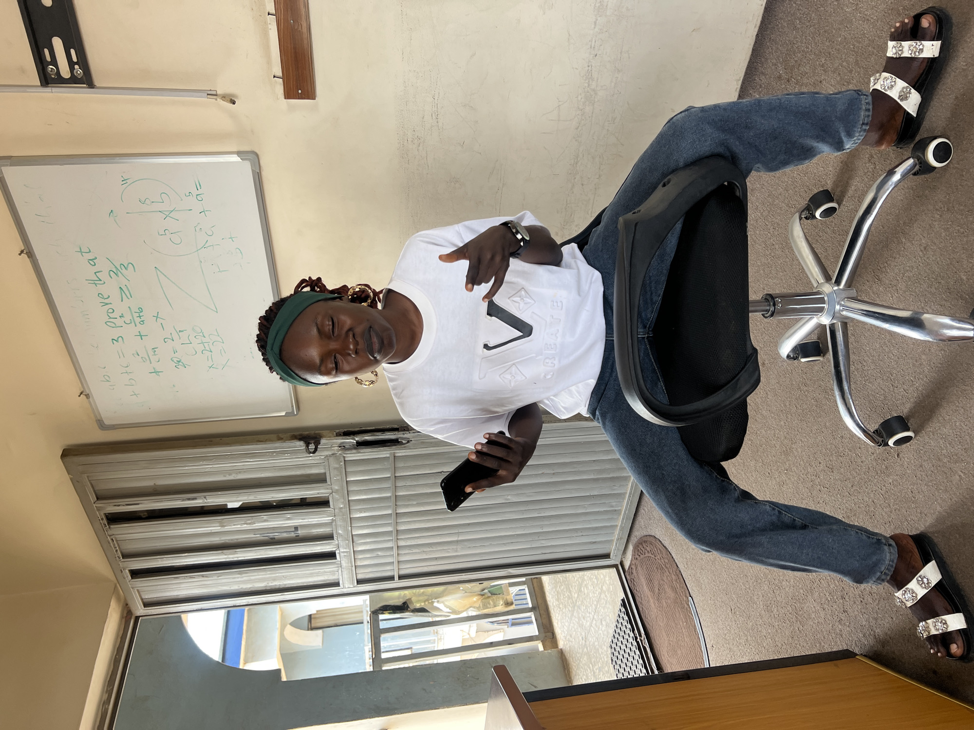 The image shows a young Black woman sitting on an office chair in a room with a whiteboard in the background, possibly a classroom or study area. The whiteboard contains mathematical equations and diagrams. The woman is wearing a white t-shirt, blue jeans, and decorative sandals. She is holding a smartphone in her left hand and gesturing with her right hand. The room appears to be located in Jos, Nigeria, judging by the architectural style of the building visible through the open doorway in the background. The doorway leads to an outdoor area with a light blue-colored building with arched windows. The floor is carpeted, and there is a wooden desk to the left of the frame.