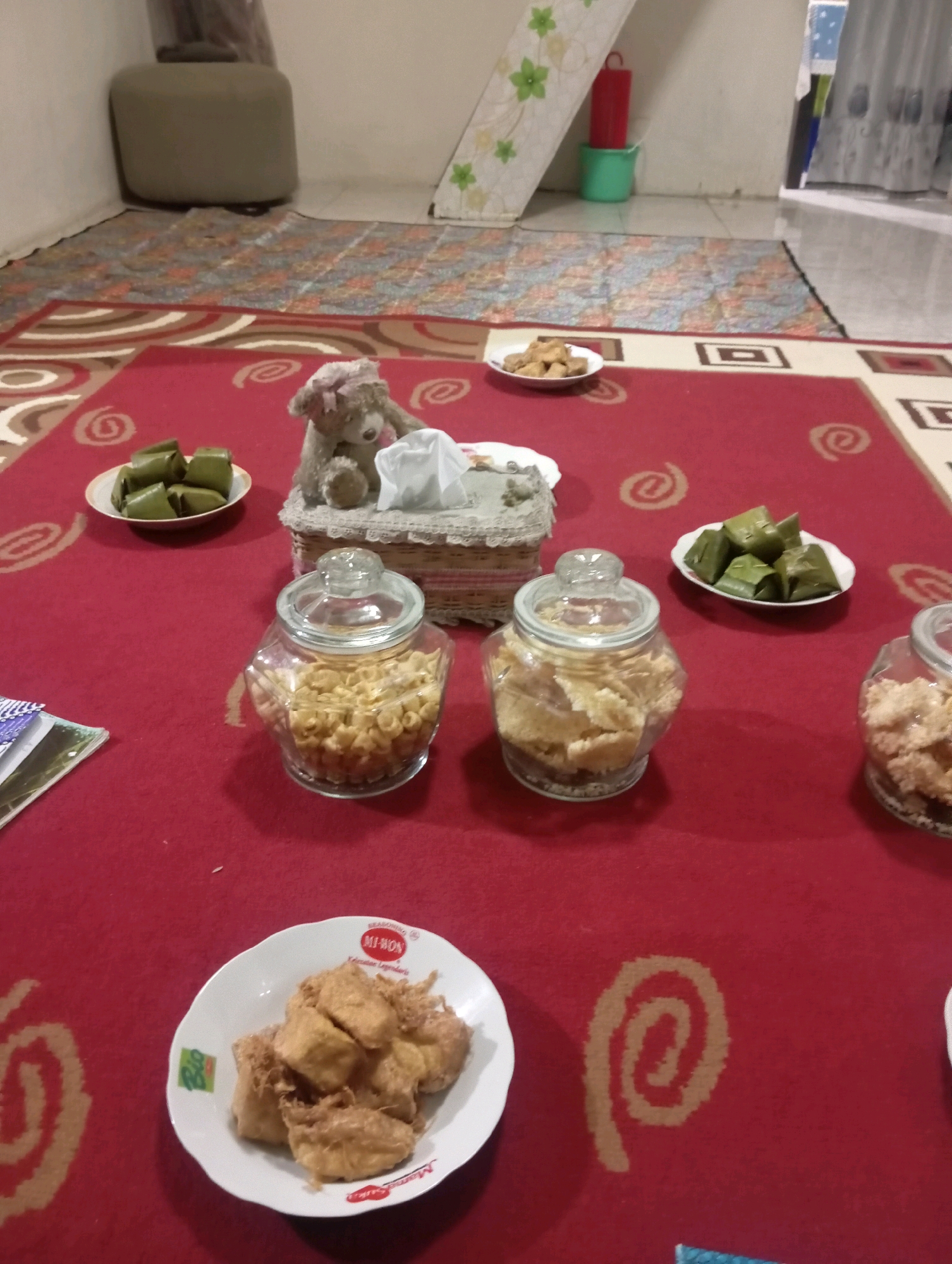 The image depicts a low-angle, close-up view of a red carpeted floor in what appears to be a room in Magelang, Indonesia. The carpet has a pattern of swirling tan and white designs. On the carpet, several plates and jars of food are arranged, suggesting a gathering or meal.
In the foreground, a white plate holds several pieces of fried tofu. To the left of this plate, a stack of magazines or papers is visible. Further back, two glass jars filled with snacks are prominently displayed. The jar on the left contains small, circular, golden-brown crunchy snacks, while the jar on the right contains larger, irregular-shaped, light-brown crunchy snacks.
Behind the jars, a white tissue box is covered with a decorative crocheted material, and a small, fuzzy teddy bear with a pink bow sits on top of it. To the left of the tissue box, a white plate holds several green, rectangular-shaped parcels, likely wrapped snacks such as lemper or nagasari.
In the mid-ground, another white plate displays a pile of golden-brown fried tofu. Further into the background, a patterned rug covers part of the floor, with a green bucket and a red cylindrical object nearby. A portion of a light-colored wall and what seems to be a piece of furniture (possibly an ottoman) are also visible in the background, along with a glimpse of a gray curtain in the far right. The lighting suggests an indoor setting, possibly in the evening or with low ambient light.