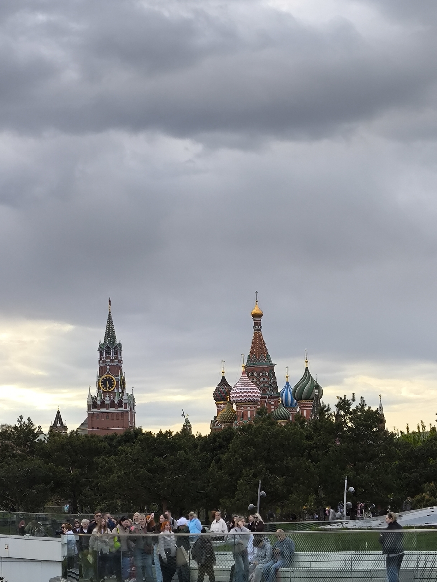 The image shows Moscow's Red Square and St. Basil's Cathedral under an overcast sky. The Spasskaya Tower of the Kremlin is visible to the left of the cathedral. A crowd of people stands on a structure in the foreground, possibly a viewing platform or bridge, looking toward the buildings. They are dressed in casual clothing, with some holding phones, likely taking photos. Trees partially obscure the base of the buildings. Street lamps can be seen between the cathedral and the people on the structure.
