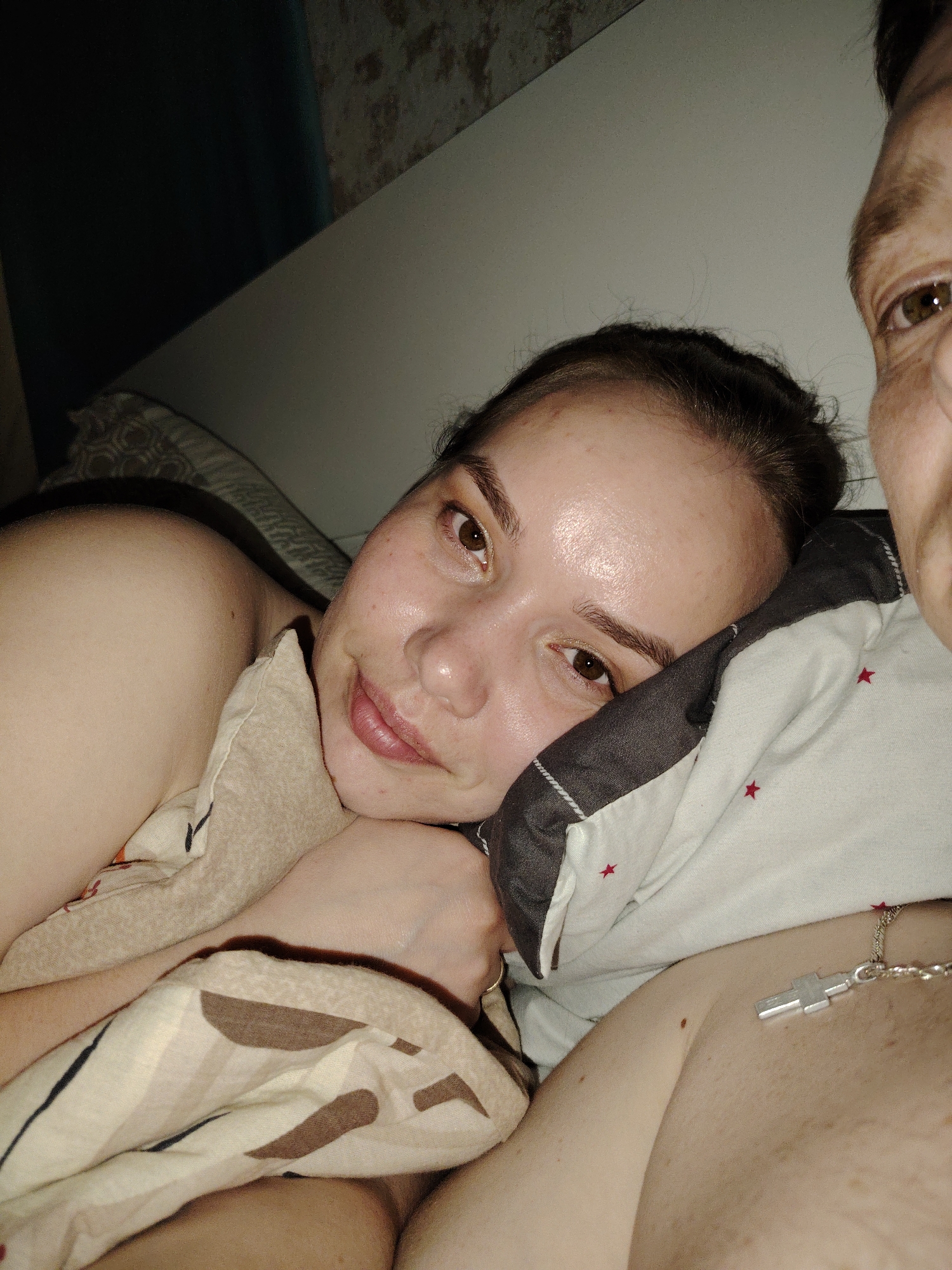 An image taken in an indoor setting, specifically a location in Марковский, Russia, features two individuals lying in bed under low light conditions. The primary subject, a female with light skin, dark hair, and brown eyes, occupies the left and central frame. She is positioned on her side, resting her head on a pillow with a light background, dark trim, and red star patterns. Her left shoulder and upper arm are exposed, and she is partially covered by a beige duvet with brown and tan abstract patterns. She is looking directly at the camera with a slight smile. To the right, a second individual is partially visible, showing their right brown eye, part of their nose, and forehead. This individual is wearing a silver-colored chain with a Chevrolet logo pendant around their neck. The background shows a dark blue/grey wall or curtain on the left and a lighter-colored wall or headboard towards the top.