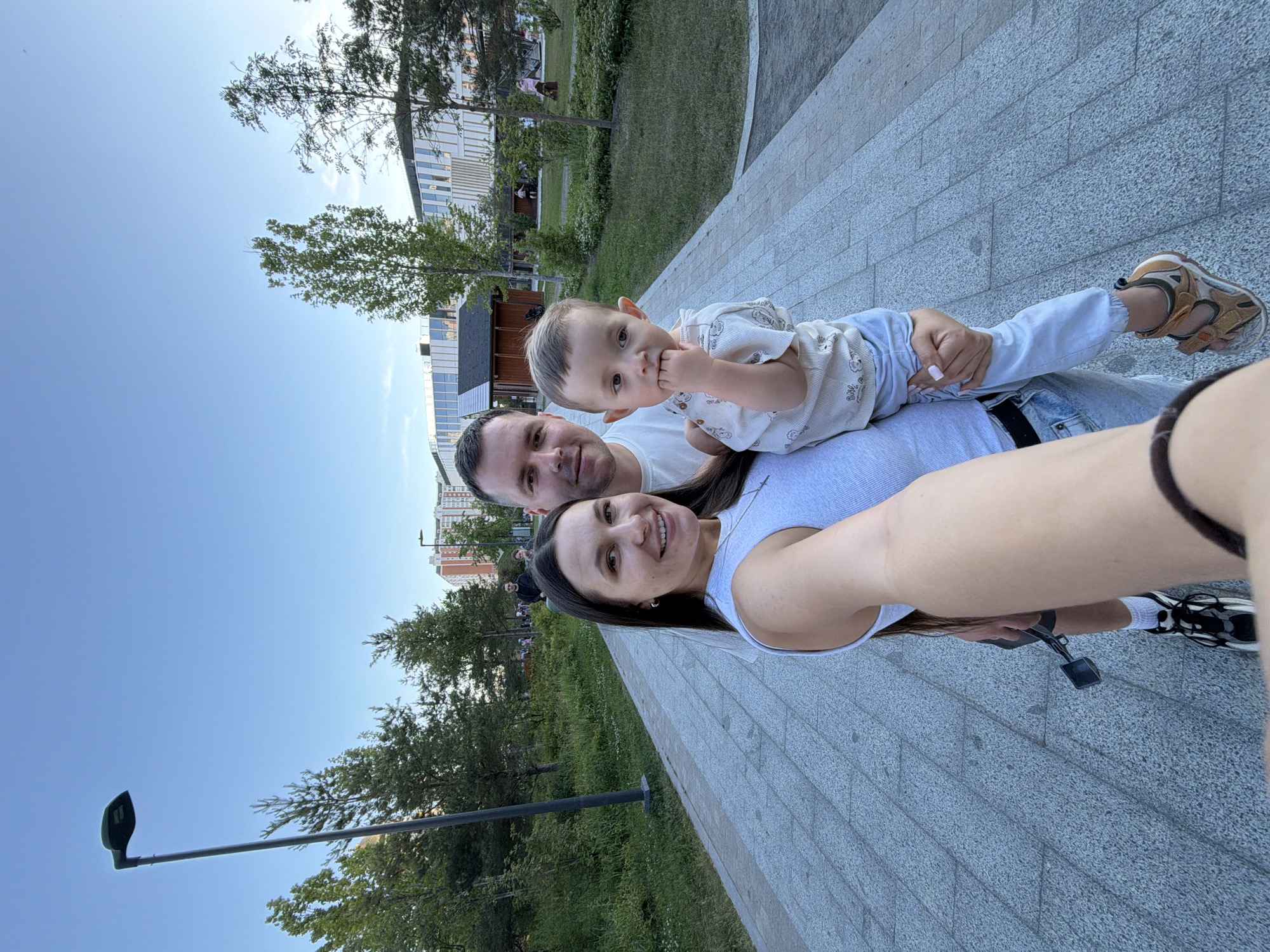 A family of three, consisting of a woman, a man, and a young child, is captured in a wide-angle selfie on a paved pedestrian walkway within a modern park environment. The woman, positioned closest to the camera and holding the child, has long dark hair, small hoop earrings, a necklace, and wears dental braces. She is smiling and dressed in a light grey ribbed tank top and light blue jeans, with white and black sneakers visible at the bottom of the frame. Black sunglasses are clipped to her waistband. The man, standing next to the woman, has short dark hair and wears a white t-shirt and light blue jeans, looking directly at the camera. The child, a toddler, is held by the woman, wears a light-colored patterned short-sleeved shirt, light blue pants, and brown and tan sandals, with one hand raised towards their mouth.
The setting is an outdoor park characterized by a wide, light grey paved pathway composed of rectangular pavers. Lush green grass and various trees, including mature conifers and deciduous trees, line the pathway. In the mid-ground behind the family, a small, dark wooden structure with a pitched roof is visible. The background features modern architecture, including a multi-story building with a distinctive tiered or wavy white facade and extensive glass windows. Further in the distance to the right, a taller residential building with orange-red and white sections can be seen. A tall, dark green streetlamp stands prominently on the left side of the frame, with another visible in the distance on the right. Several other individuals are visible as blurred figures in the background, indicating a public space. The sky above is clear and light blue, suggesting daytime.
This scene is located within the **Skolkovo Innovation Center** in Moscow, Russia, identifiable by the distinctive modern architecture integrated with contemporary park infrastructure.
