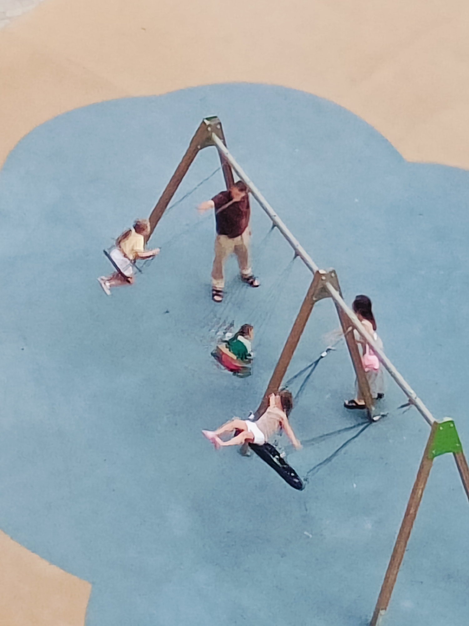 This is an aerial, slightly blurry, full-color photograph of a playground. The ground is a pale blue surface with a lighter beige, abstract shape in the upper left corner. Several children and one adult are visible interacting with a swing set.
The swing set has a wooden A-frame structure with metal supports. There are at least three swings. On one swing, a child in a yellow shirt and white shorts is swinging. Nearby, an adult in a maroon shirt and khaki pants stands with arms outstretched. Another swing is occupied by a child wearing a green and white outfit. A third swing is being used by a child in a white top and white bottoms, lying on a black swing seat. A child with dark hair and a pink bag is standing near the base of the swing set.
The scene depicts a playful and active moment. The lighting suggests it is daytime, likely sunny, with no indication of specific weather conditions. The location is identified as Krasnodar, Russia, though no specific landmarks are visible to confirm this. The emotions conveyed are primarily joy and activity, typical of a playground setting. No text is visible in the image.