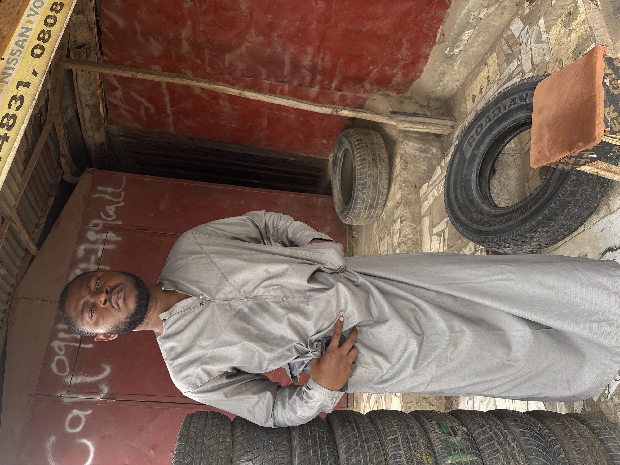 A man with a dark beard and mustache stands and looks directly at the camera in what appears to be a tire shop or an informal market stall. He wears a light grey, long-sleeved tunic, with his left hand in his pocket and his right hand holding a dark-colored smartphone.
Behind him is a red corrugated metal surface, possibly a door or wall, with white graffiti that clearly spells "Call". To the man's left, a neat stack of used tires is visible on the ground. To his right, two single tires hang against a textured concrete wall, with the brand name "ROADIAN" visible on the tire closer to the foreground. Above these hanging tires, an orange-brown piece of fabric is draped over a black and gold patterned object.
In the upper left corner of the frame, part of a yellow sign is visible, displaying text that reads "NISSAN VO" and phone numbers "4831, 0808". Wooden beams and other rustic construction materials are also noticeable, suggesting a functional, semi-outdoor setting. The scene is illuminated by natural daylight. The overall image captures a candid moment in a commercial area, consistent with a street-side business in Lugbe, Nigeria.