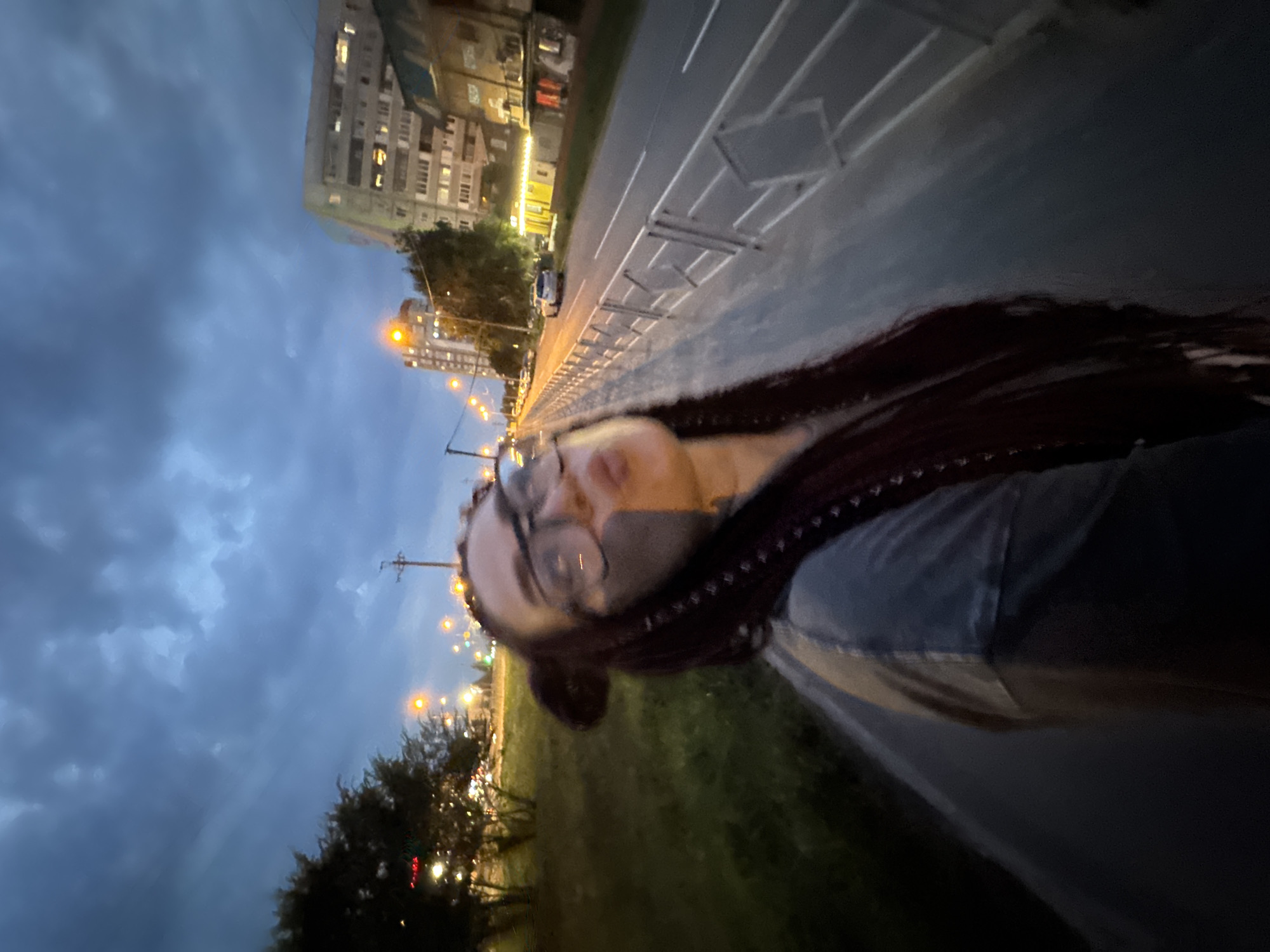 A young woman with braided hair and glasses poses for a selfie outdoors at dusk in Tolyatti, Russia. She is wearing a grey t-shirt with a dark stripe down the left side of her torso. Her face is partially obscured by her hand and is blurred due to the camera's close proximity. She has dark, smudged makeup around her eyes.
The background shows a street scene with a sidewalk, a metal fence, and a road with a single white car visible. On the right side of the road, there are several multi-story apartment buildings, some with illuminated windows. Streetlights are visible along the road, casting a yellow glow. Trees line the left side of the sidewalk, with red and yellow lights from nearby buildings or traffic visible through the foliage. The sky is a dark, cloudy blue, indicating it is either twilight or a stormy evening. The image was likely taken from a wide-angle perspective, creating a fisheye effect.