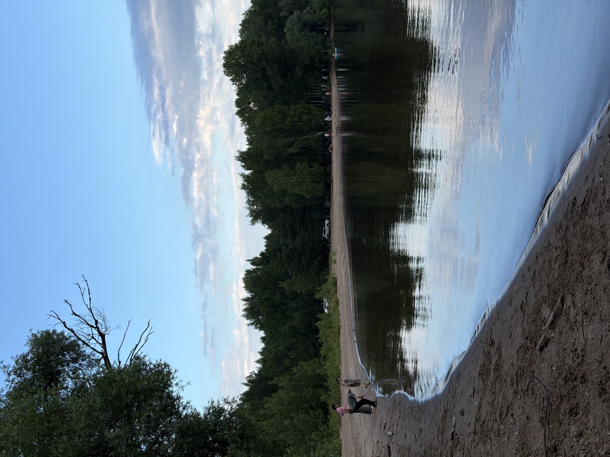 In Noviy, Russia, this image captures a serene scene at a lakeside beach. The foreground is dominated by a sandy shore with sparse debris. To the left, three individuals are present. One person, wearing a pink top, stands facing left. Beside them, another person in a dark green shirt and black pants appears to be interacting with the sand near the water's edge. A third person, in a patterned dress, stands a bit further back.
The water, dark and reflective, gently laps at the shore, mirroring the sky and the treeline. Across the water, a dense forest of green trees forms the backdrop. A few vehicles are visible parked along the sandy area behind the treeline. In the distance, to the right, a person is seen on a blue float in the water. The sky above is a gradient of pale blue, transitioning from lighter shades near the horizon to a deeper hue overhead, with scattered white and gray clouds. The overall atmosphere is tranquil, suggesting a quiet day by the lake.