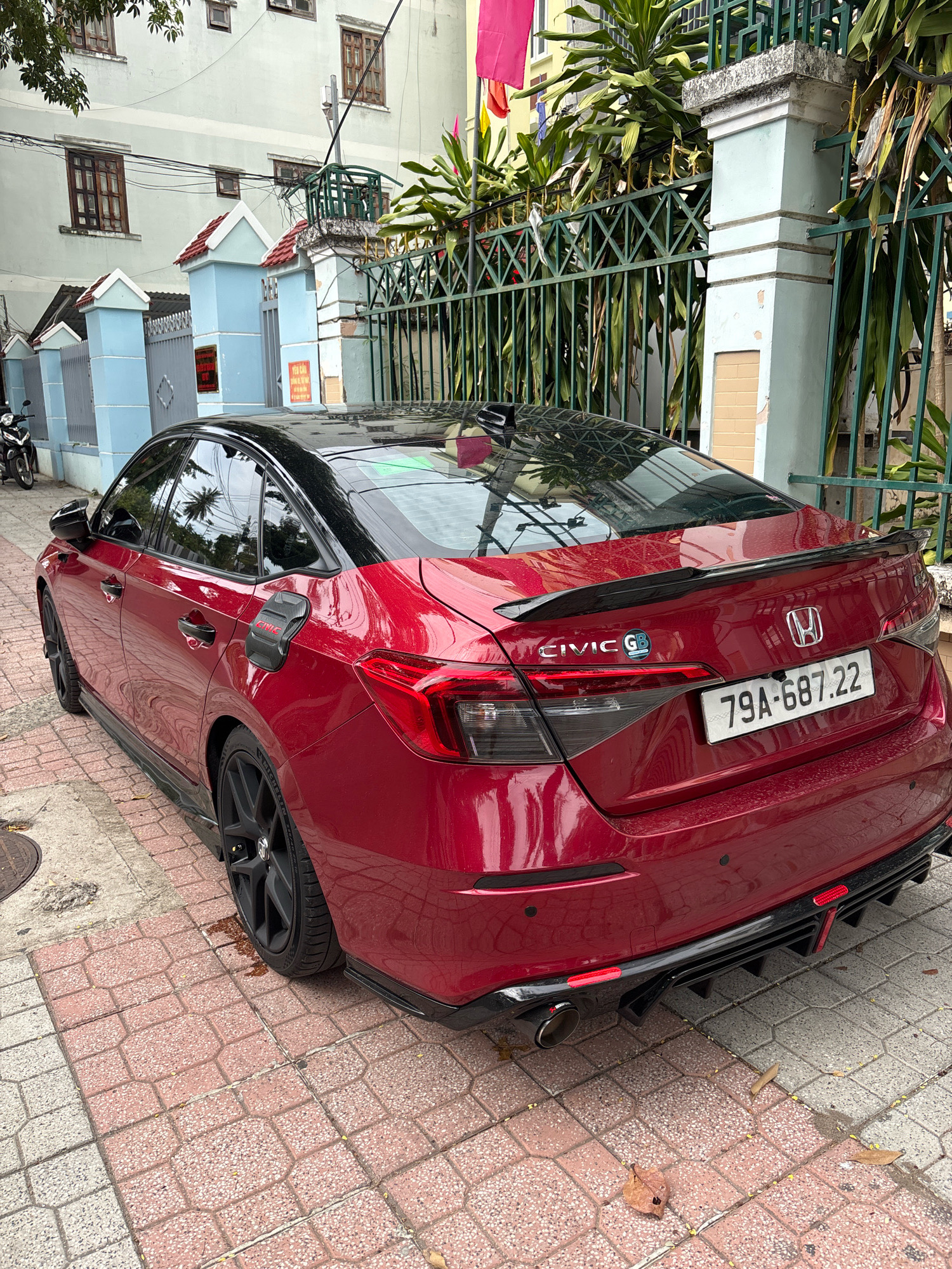A red 2022 Honda Civic G sedan is parked on a sidewalk paved with red and white interlocking bricks in Nha Trang, Vietnam. The car has a black roof and spoiler. Its rear features a license plate reading "79A-687.22", a "CIVIC G" emblem, and a Honda logo. The car is positioned on the right side of the frame, angled away from the viewer. Behind the car is a green metal fence with decorative gold elements and light blue pillars topped with red-tiled roofs. A building with light gray walls and dark-framed windows is visible in the background, along with lush green foliage. The sky is overcast.