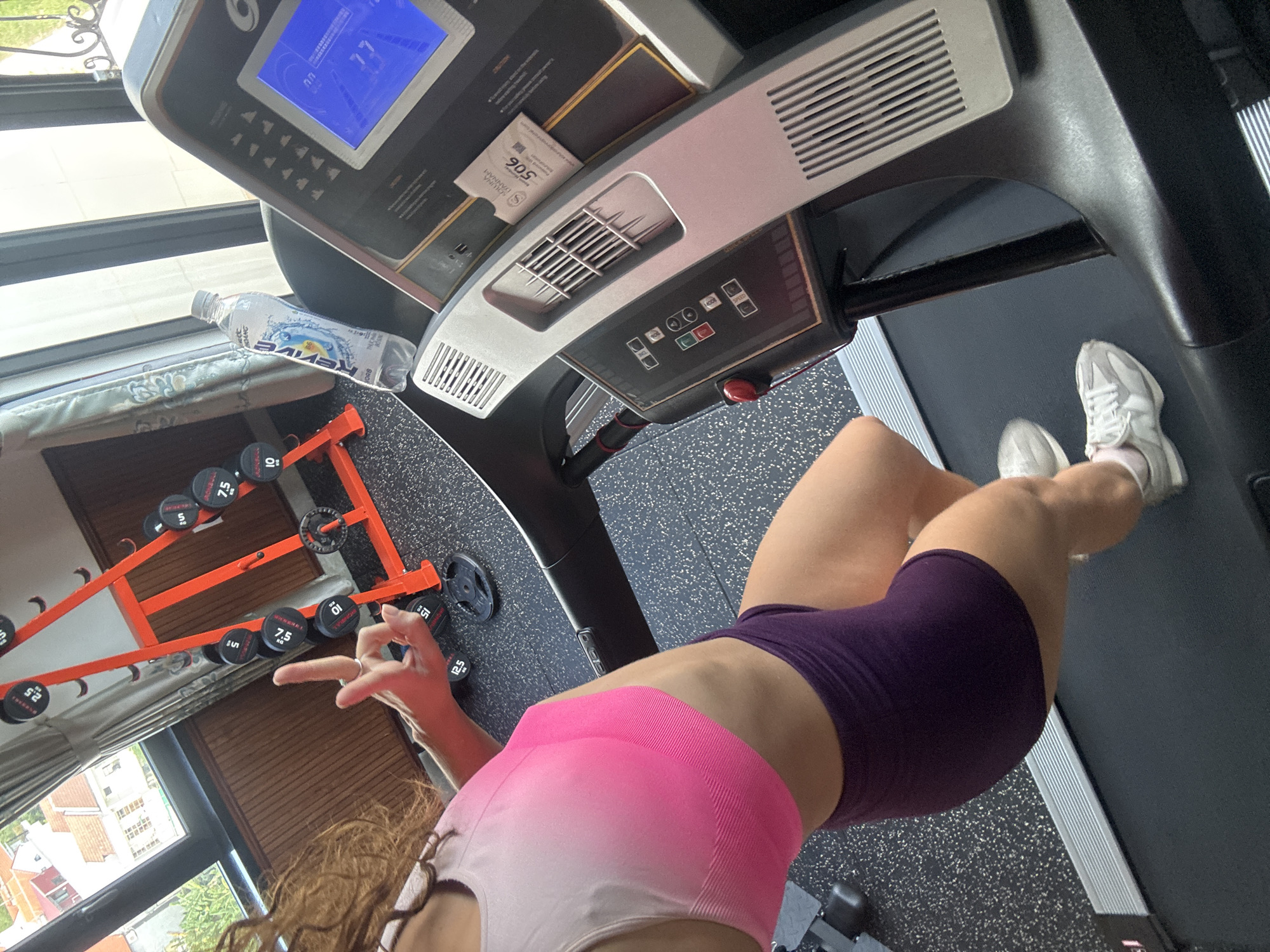 A high-angle shot depicts a person in workout attire standing on a treadmill, likely in a home gym environment. The person is wearing a pink and white sports bra and purple exercise shorts with white sneakers. The treadmill features a digital display showing "77" and control panels. A bottle of "Revive" water rests on the treadmill's console. To the left of the treadmill, a rack holds an assortment of dumbbells, ranging from 5 to 12.5 units (likely pounds or kilograms). In the background, a window reveals a view of buildings and greenery, indicative of an urban environment. Based on the architectural style visible through the window and the "Revive" water brand, it's possible the location is in Southeast Asia. It is specified to be in Hội An Ward, Vietnam. The person is making a hand gesture with their left hand.
