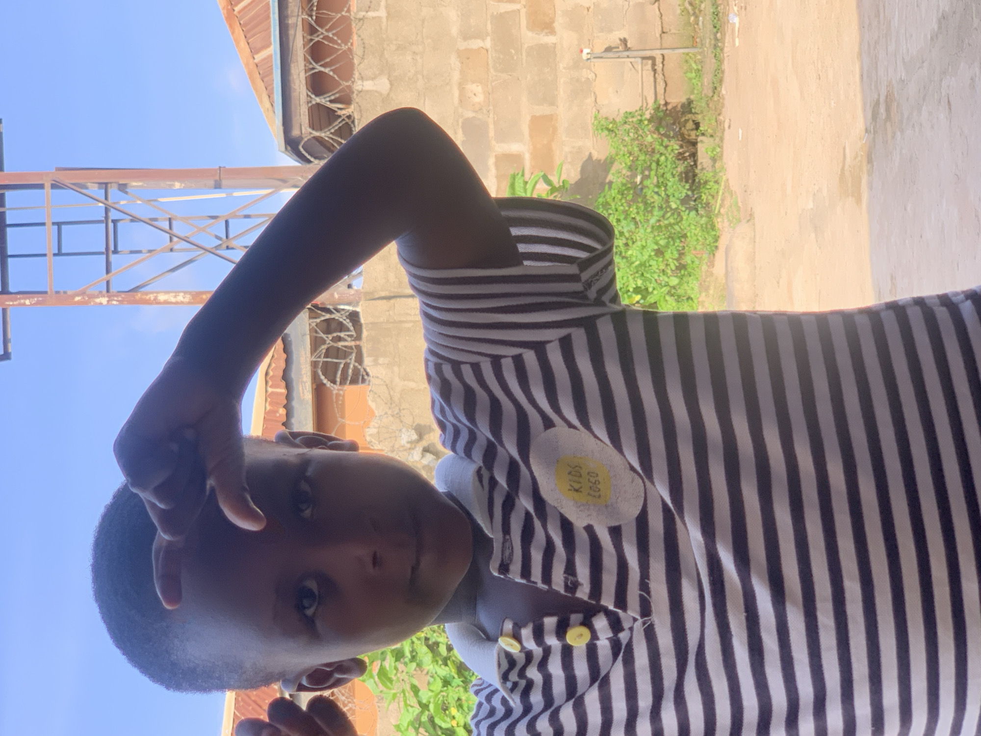 A young child, appearing to be of African descent, is pictured from the chest up, looking directly at the camera with a neutral expression. The child has short, dark hair and is wearing a black and white striped polo shirt with yellow buttons. A circular patch with yellow text is visible on the left side of the shirt, appearing to read "KIDS LOGO". The child's right hand is raised, with fingers forming a gesture near their forehead.
The background consists of a brick wall under a bright blue sky, with some green foliage to the left and a portion of a metal structure on the right. The overall scene suggests an outdoor setting, possibly a residential area in Sabon Tasha, Nigeria, with the characteristic brick construction and dry, dusty ground often seen in such locations. The lighting indicates it is daytime.