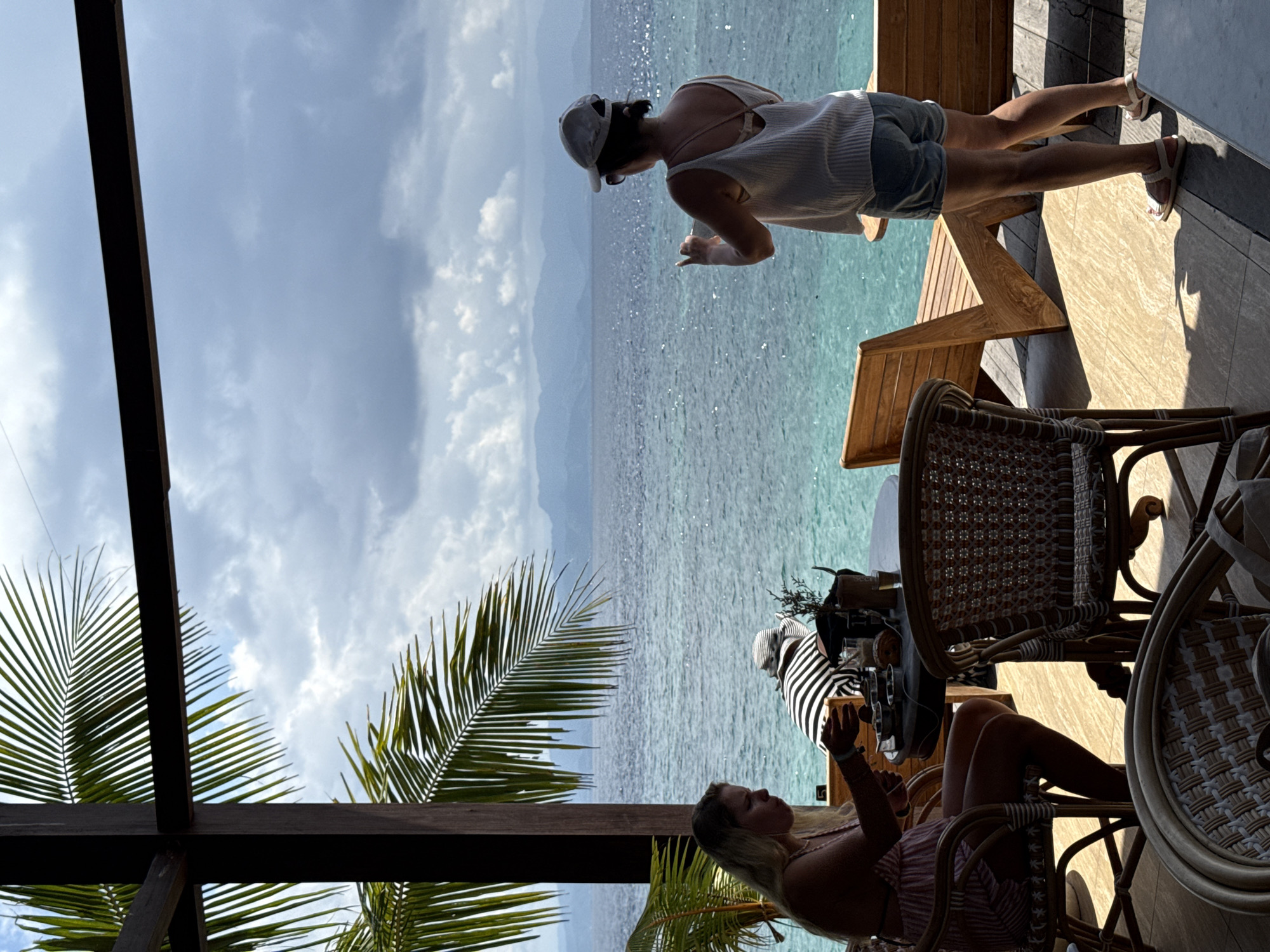 The scene is captured at a restaurant, likely in the coastal city of Gianyar, Indonesia, given the presence of a tropical beach environment. The focus is on a woman with a baseball cap leaning over the edge of a wooden deck, presumably looking at the ocean. She wears a light top and denim shorts with sandals. There is a wooden table and a wooden A-frame chair next to her. Seated below her, partly obscured by chairs, is another woman with long blond hair. Palm trees are visible in the foreground. The ocean is visible and there are islands in the distance.