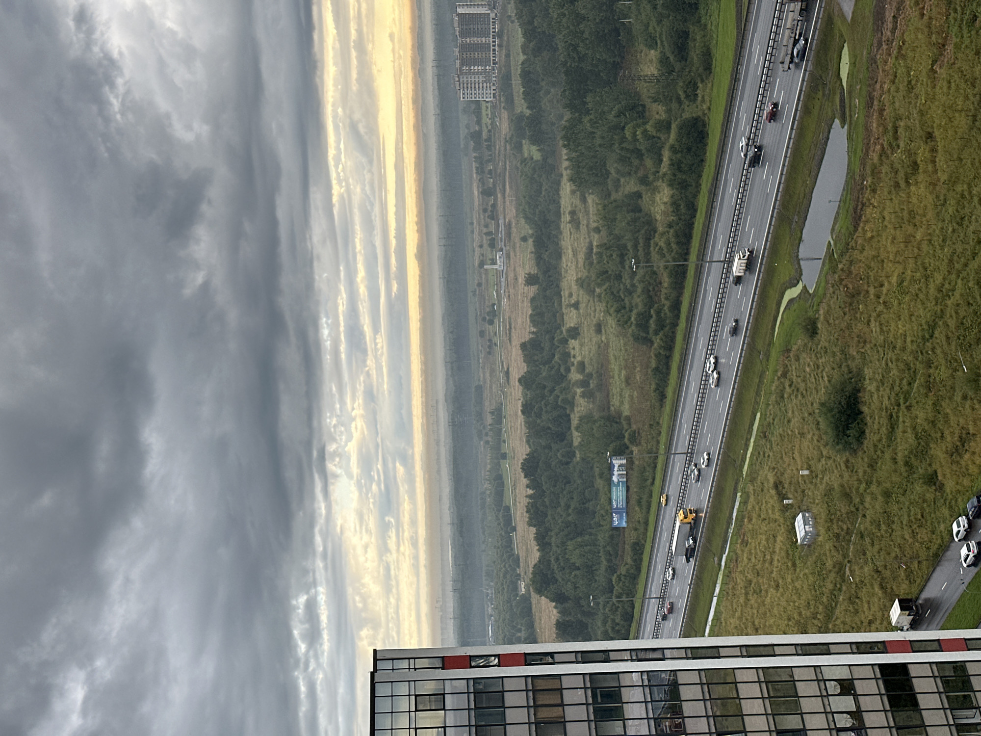 The image shows an aerial view of a highway passing through a green, sparsely populated area on the outskirts of Kudrovo, Russia. The sky is overcast with dark, dramatic clouds. To the left, a modern apartment building with a glass facade is partially visible. The highway, wet from recent rain, is active with various vehicles, including cars, a yellow truck, and a trailer truck. Beyond the highway, the landscape is dominated by fields and dense green trees. In the distance, the faint outlines of industrial structures and apartment complexes can be seen, suggesting the proximity of urban development. A small pond or waterlogged area is present next to the highway, and a small shed-like structure is situated in a grassy field. The overall impression is one of a developing urban fringe where nature meets infrastructure.