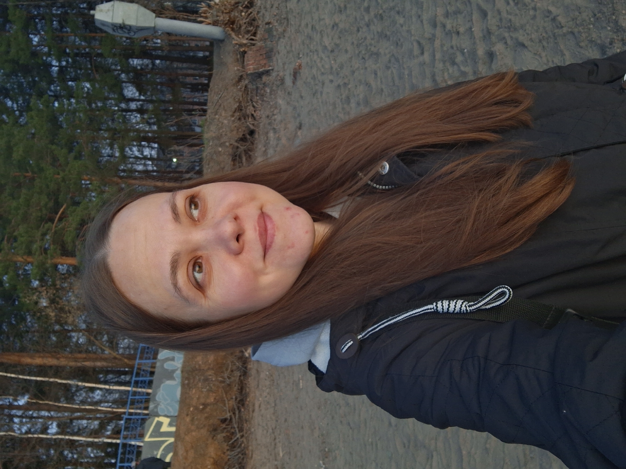 A young woman with long brown hair is taking a selfie. She has fair skin and light brown eyes, and is wearing a black jacket with white and black trim. The background shows a sandy area with trees, a light pole, and a blue fence. Graffiti is visible on a wall in the distance. The photograph was taken outdoors. The location is the city of Chaikovsky, Russia.