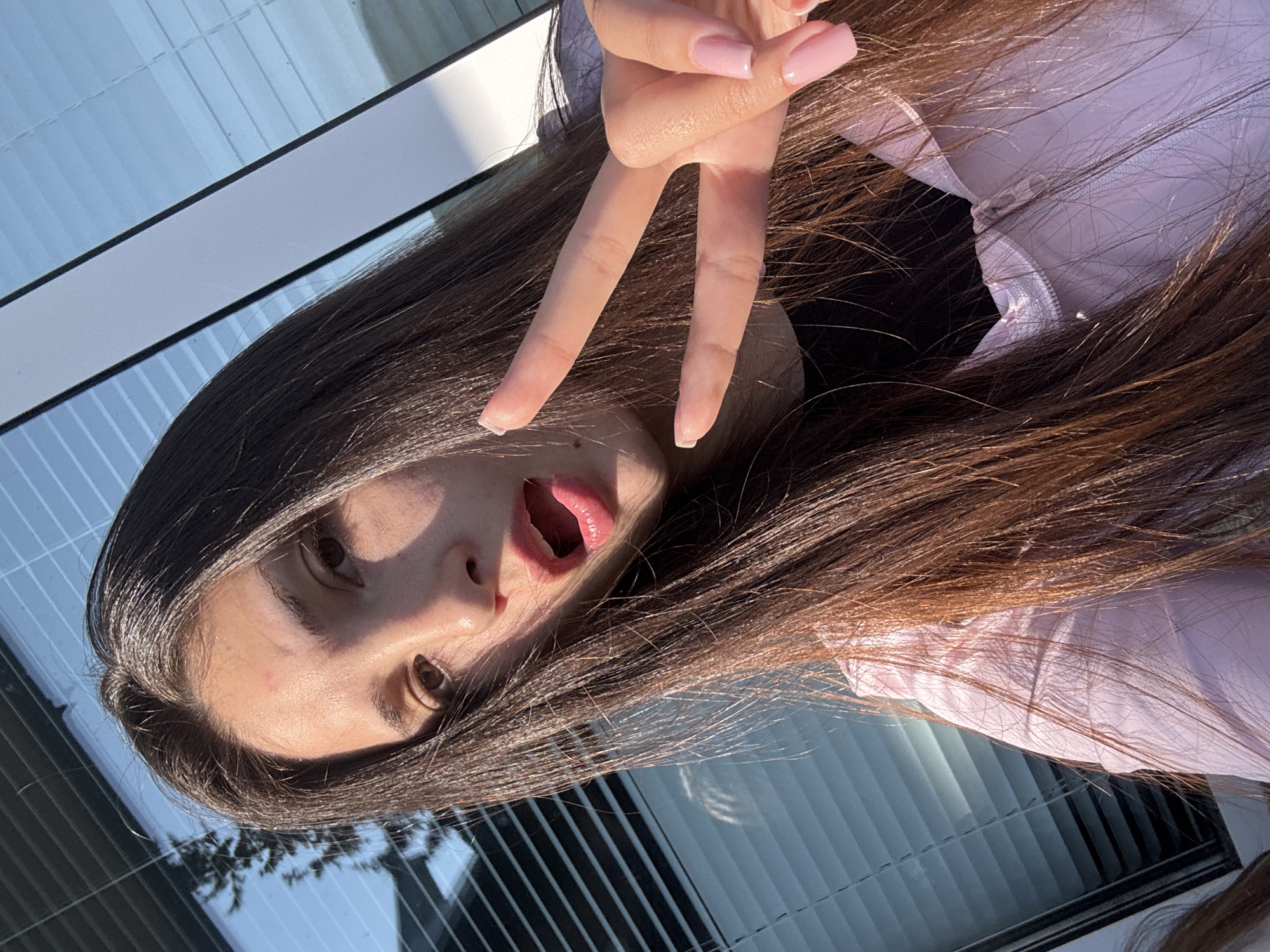 A young woman with long, brown hair is taking a selfie outdoors. She is wearing a light purple top and has light pink acrylic nails. Her mouth is open, and she is making a peace sign with her right hand, her fingers extended. Her eyes are open, and she appears to be looking at the camera. Sunlight is illuminating her face from the right, casting shadows and highlighting her features. The background is a reflection of what appears to be a modern building with large windows and horizontal blinds. The scene suggests a sunny day, likely in the afternoon, given the angle of the light. The overall mood is casual and playful. No text is visible in the image.