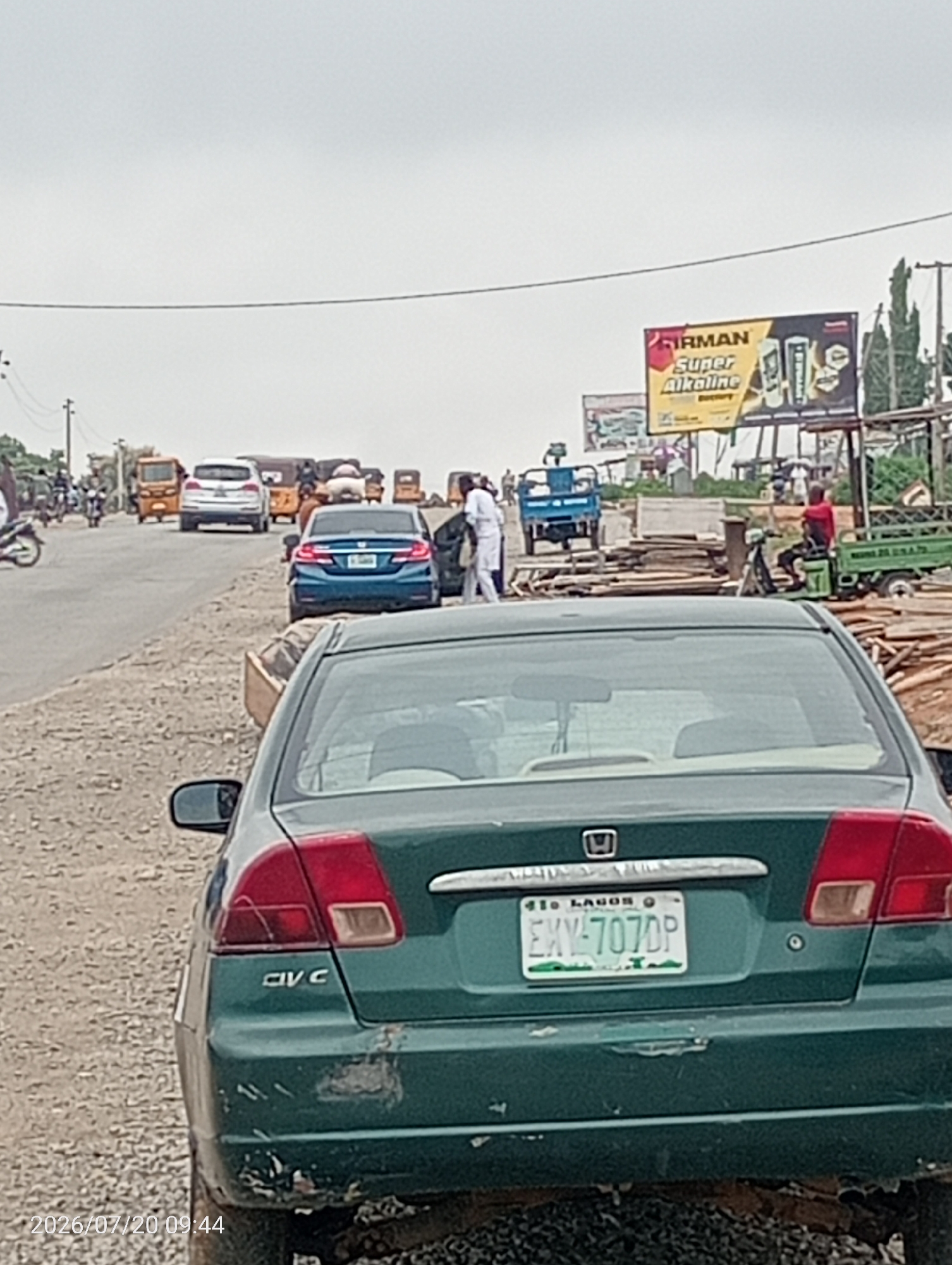 The image shows a street scene in Suleja, Nigeria, with several vehicles and people present. The foreground is dominated by the rear of a green Honda Civic car, displaying a license plate that reads "EKY-707DP". The car shows signs of wear and tear, with damage to its bumper. The street itself is unpaved and appears to be under construction or in a state of disrepair, with piles of wood and building materials visible along the roadside.
Further down the street, a blue sedan is parked, and other vehicles, including motorcycles and a small truck, are present in the background. A billboard advertising "FIRMAN Super Alkaline Battery" is visible in the distance. Several individuals are seen, some standing on the roadside and others operating vehicles. The sky is overcast, suggesting a cloudy day. The timestamp in the lower left corner indicates the image was taken on July 20, 2026, at 09:44.