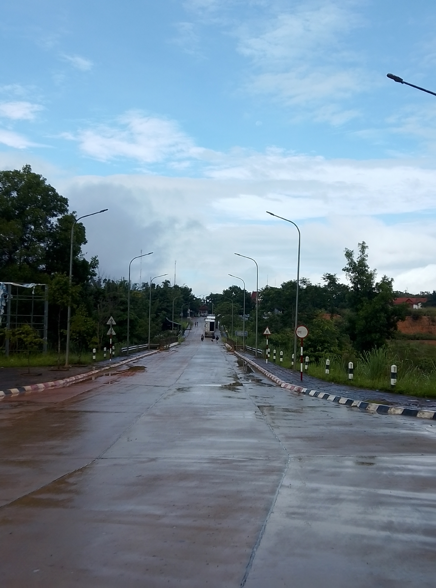 The image displays a straight road extending into the distance under an overcast sky with visible patches of blue. The road surface, appearing to be concrete, is wet and reflective from recent rain, showing puddles and a sheen. Tall streetlights line both sides of the road, receding towards the horizon.
On the left, a curb with red and white striped markings borders the road, followed by a metal frame structure and dense green trees. Further along, black and white striped bollards mark the roadside. The right side of the road also features a curb with black and white striped markings and similar bollards. Beyond these, a grassy bank slopes upward, leading to distant buildings with reddish roofs partially obscured by trees. Two road signs are visible on the right: a triangular warning sign and a circular sign with a red border and white horizontal bar, commonly indicating "no entry" or "no stopping."
In the far distance, a white truck or large vehicle is visible on the road, accompanied by a few small human figures. The scene suggests a daytime setting, likely after a period of rain, giving the environment a damp and serene atmosphere. The presence of streetlights and road markings indicates a structured roadway within a landscape that blends natural greenery with some developed areas. No specific text is legible on the visible signs or objects.