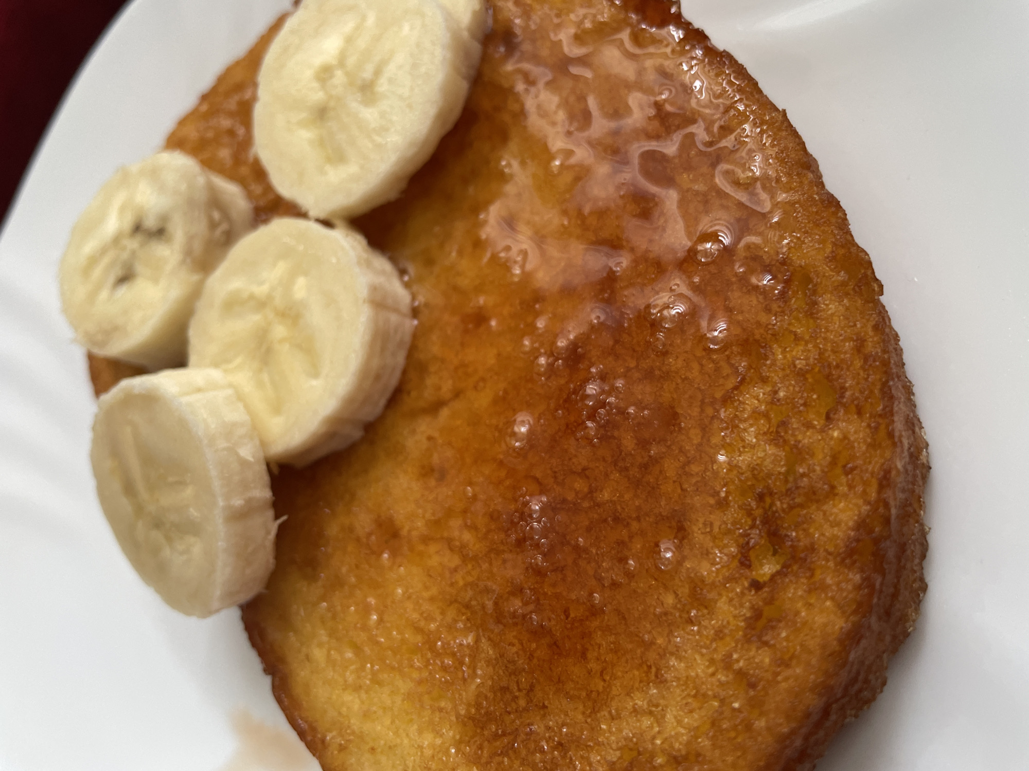 A close-up shot shows a pancake on a white plate topped with banana slices and syrup. The pancake appears to be golden brown and glistening with syrup. Four banana slices are placed on the left side of the pancake. The plate is plain white. The location is not discernible from the provided image.