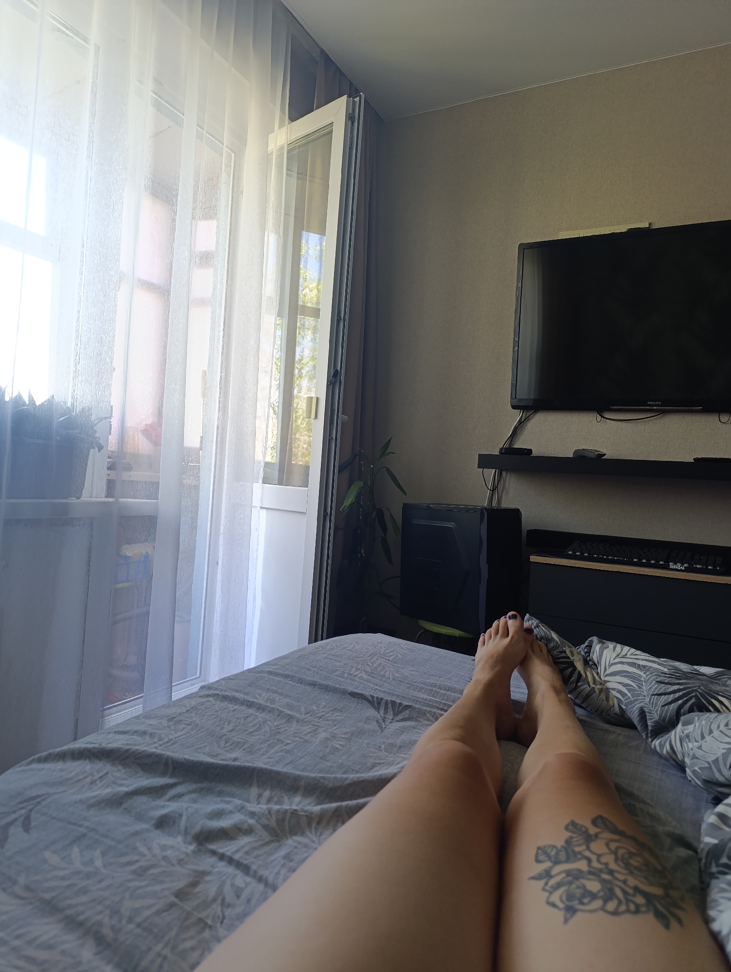 The image depicts a person's legs and feet resting on a bed with a grey, leafy-patterned duvet. The person has dark nail polish on their toes and a floral tattoo on their right thigh. The bed is positioned in a room where sheer white curtains are drawn over a window or balcony door, allowing light to enter. Beyond the curtains, trees and a building are visible. To the right of the bed, a dark television is mounted on the wall above a shelf containing a remote control and other electronic devices. Below the shelf, a black computer tower sits next to a dark cabinet with a keyboard on top. The walls of the room are a neutral, textured color.