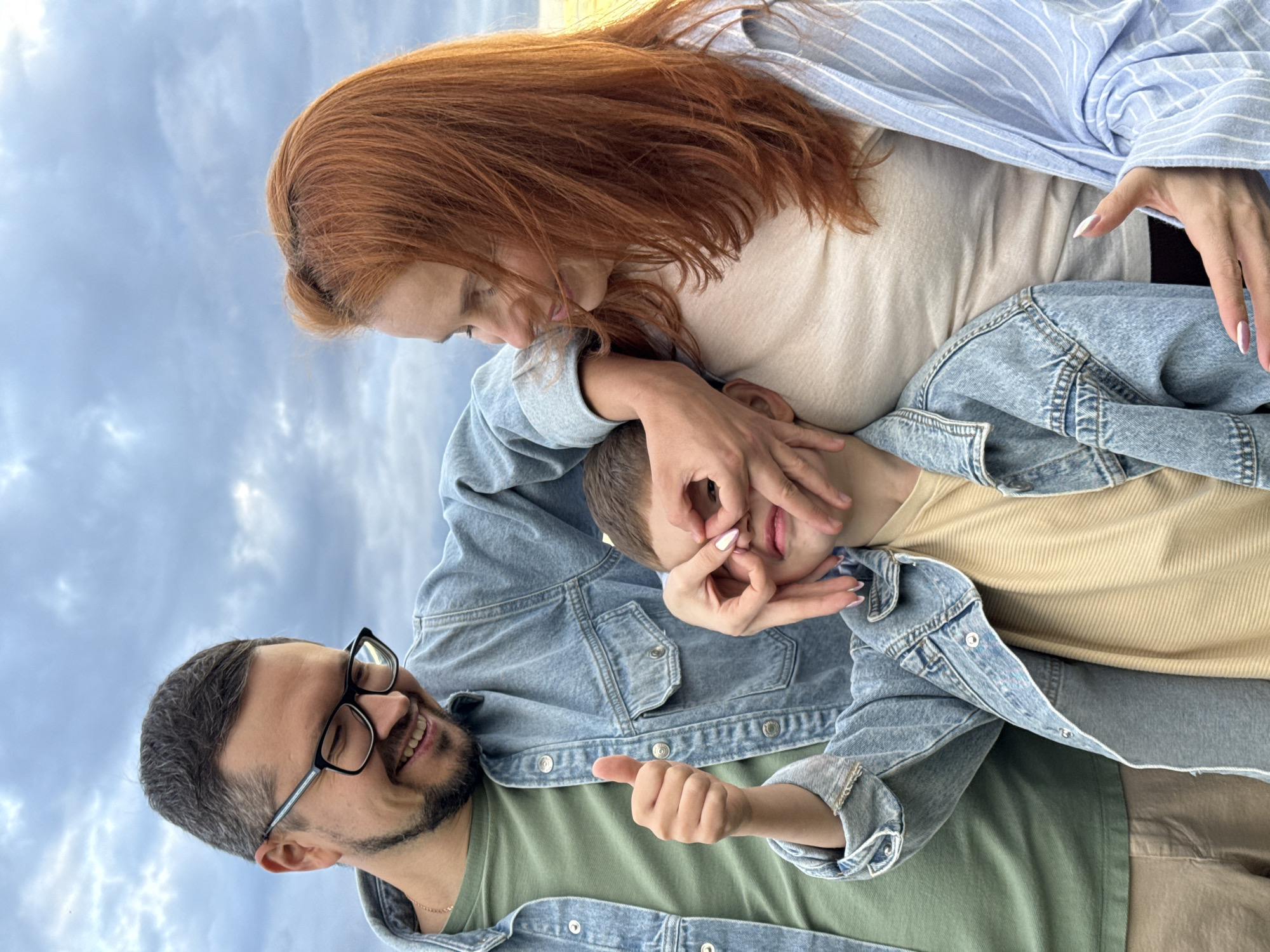 A family, consisting of a man, a woman, and a young boy, are posing for a photo outdoors under a cloudy sky. The man, on the left, wears glasses, a green t-shirt, and a denim jacket. He is smiling and giving a thumbs-up. The woman, on the right, has red hair and is wearing a beige top and a light blue striped shirt over a denim jacket. Her hands are touching the boy's face, playfully covering his eyes. The boy is in the center, wearing a yellow and beige striped shirt and a denim jacket, with his eyes covered by his mother's hands. The background appears to be a field, possibly on the outskirts of Olkhovka, Russia, given the natural, open landscape and the overcast weather.