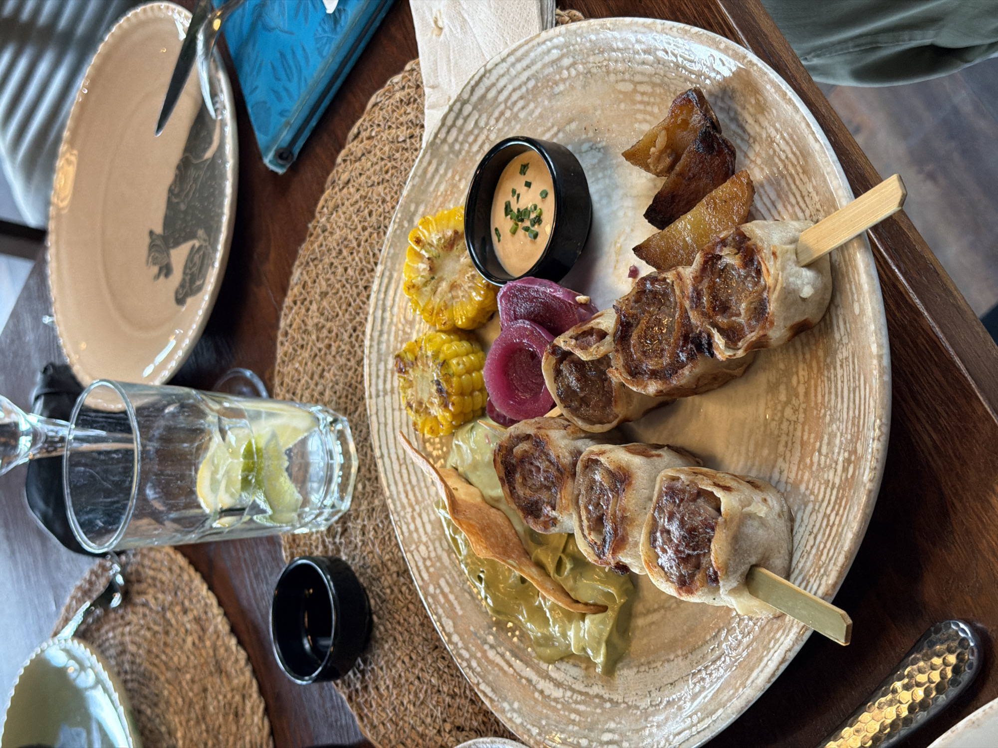 A wooden table holds a beige plate with two skewers of meat rolls, grilled corn, pickled onions, fried potatoes, and a sauce in a small black bowl. A glass of water with lemon, an empty beige plate, and silverware are also on the table. The scene appears to be at a restaurant. No individuals are clearly visible in the frame to confirm the location in Varna, Bulgaria.