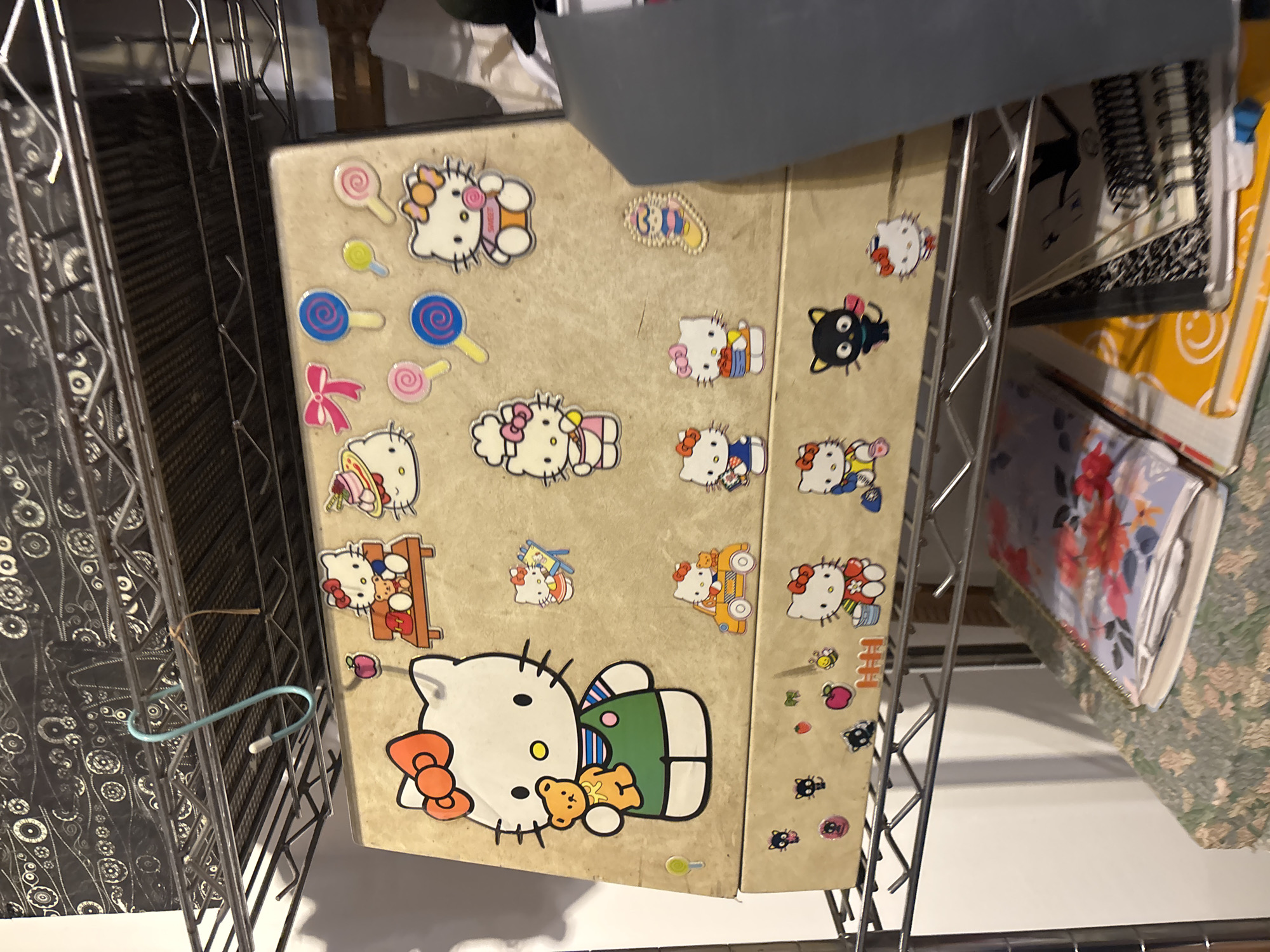 The image shows a "Hello Kitty" themed binder stored on a wire shelf. The binder is beige with various "Hello Kitty" stickers and is positioned among other items on the shelf. The background reveals the wire shelving unit and other books and binders. The location cannot be determined from the image.