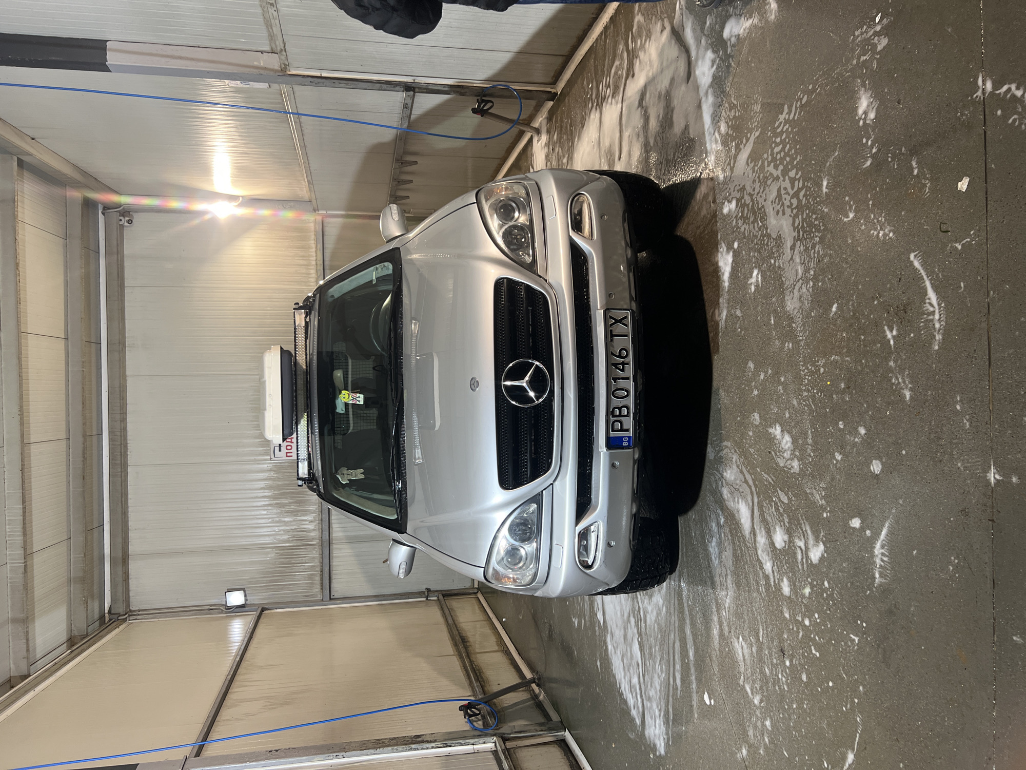 The image shows a silver Mercedes-Benz SUV being washed in an indoor car wash facility. The car is positioned facing the camera. There's a roof rack on the car. The floor around the car is wet with soap residue. A blue hose and a pressure washer wand are visible. The location is potentially in Asenovgrad, Bulgaria, as indicated by the car's license plate "PB 0146 TX" which is registered in the Plovdiv Province, where Asenovgrad is located. The interior of the car wash is constructed with corrugated metal walls and overhead lighting. Part of a person and their clothing is visible at the top of the image.