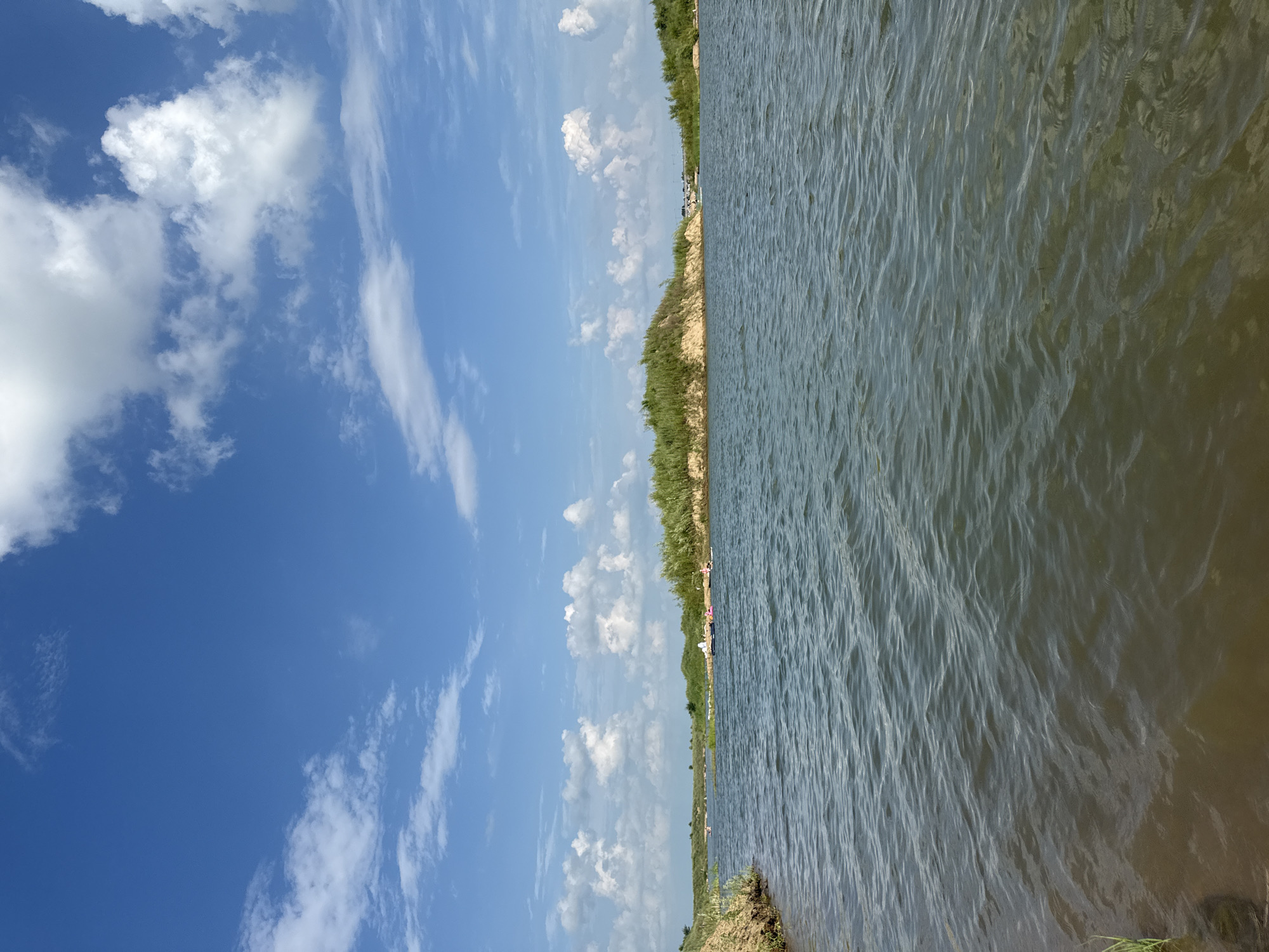 The image shows a body of water with ripples and a few small boats on the surface. In the background, there is a shoreline with green vegetation and a sandy embankment. The sky is blue with scattered white clouds. The scene appears to be a natural landscape, possibly a lake or river, under a bright, partly cloudy sky. The location is identified as Blagoveshchensk, Russia.