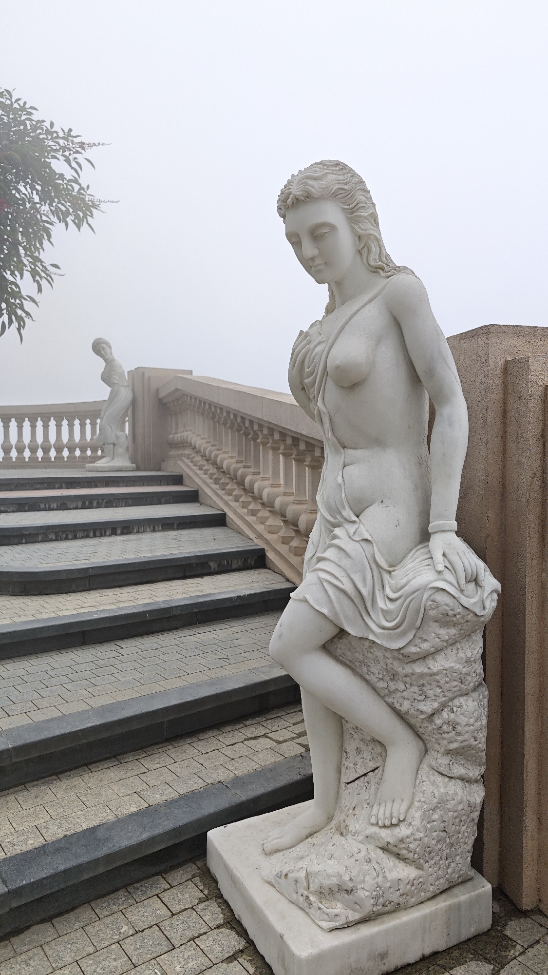 The image shows a marble statue of a woman in a classical style. She is leaning on a stone pedestal. In the background is a staircase with a balustrade, and another statue can be seen in the distance. The scene is shrouded in fog. The statue is located in Đà Nẵng, Vietnam, at the Ba Na Hills complex.