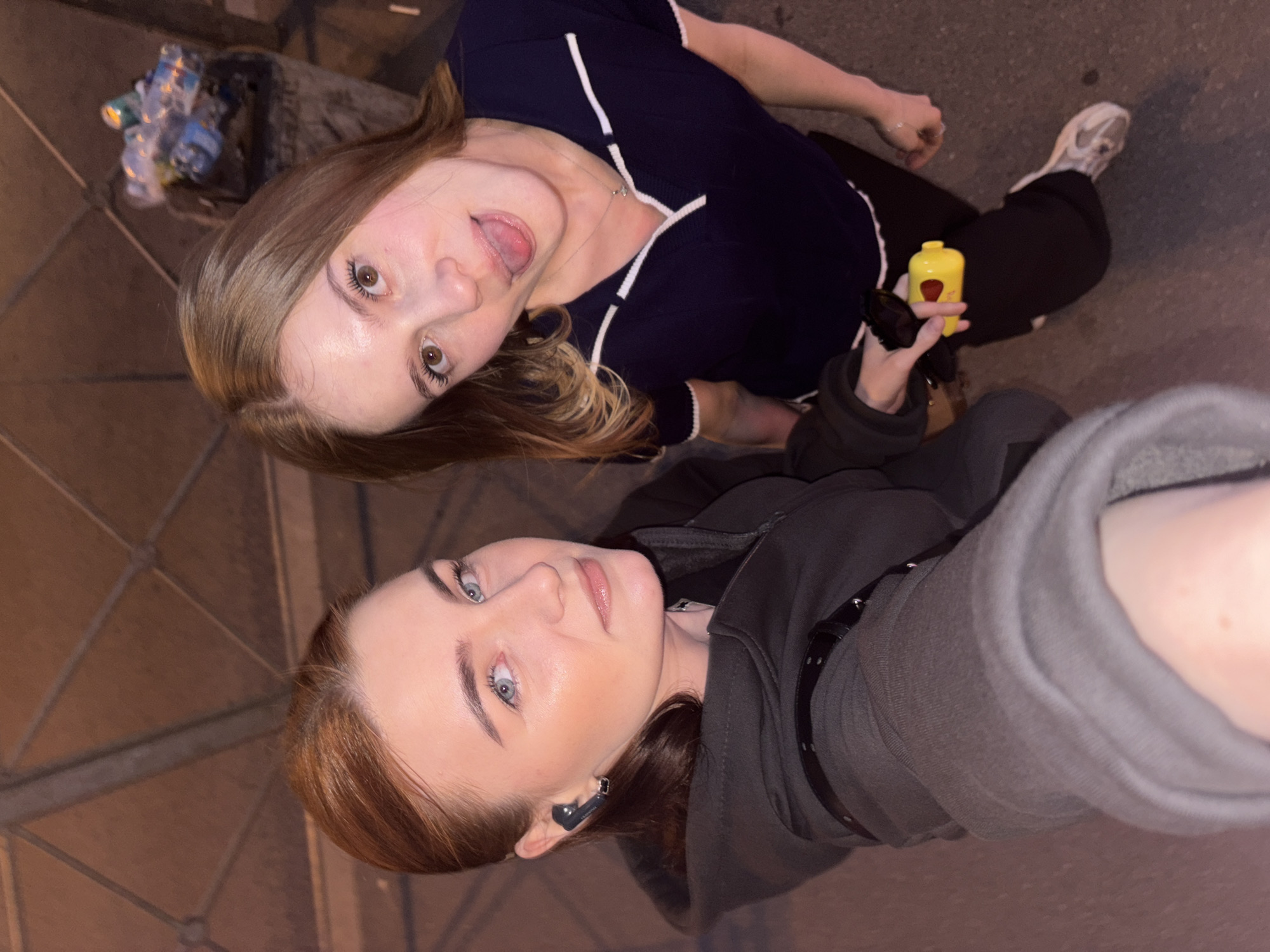 Two young adult women are captured in a selfie-style photograph in the city of Saint Petersburg, Russia, under dim artificial lighting, possibly outdoors at night or in a covered urban area.
The woman on the left, positioned closer to the camera, has auburn hair pulled back with a center part, blue eyes, and fair skin. She wears a dark grey hooded sweatshirt over a darker top and a black studded strap across her chest. A black in-ear monitor is visible in her left ear, accompanied by a small silver earring. Her expression is neutral as she looks directly at the camera.
The woman on the right, positioned further back and crouching or kneeling, has long brown hair and brown eyes. She wears a dark blue short-sleeved top with white trim on the V-neck collar and sleeves, black pants, and light-colored sneakers. She has a thin silver necklace with a small pendant. Her expression is playful, with her tongue extended towards the camera. She holds a small, bright yellow, cylindrical object with a black top in her left hand.
The ground is dark asphalt or concrete. In the background, above the subjects, a dark, geometric grid pattern is visible on what appears to be a ceiling or overhead structure. To the right, behind the second woman, a dark, cylindrical trash receptacle is present, filled with discarded plastic bottles and other refuse.