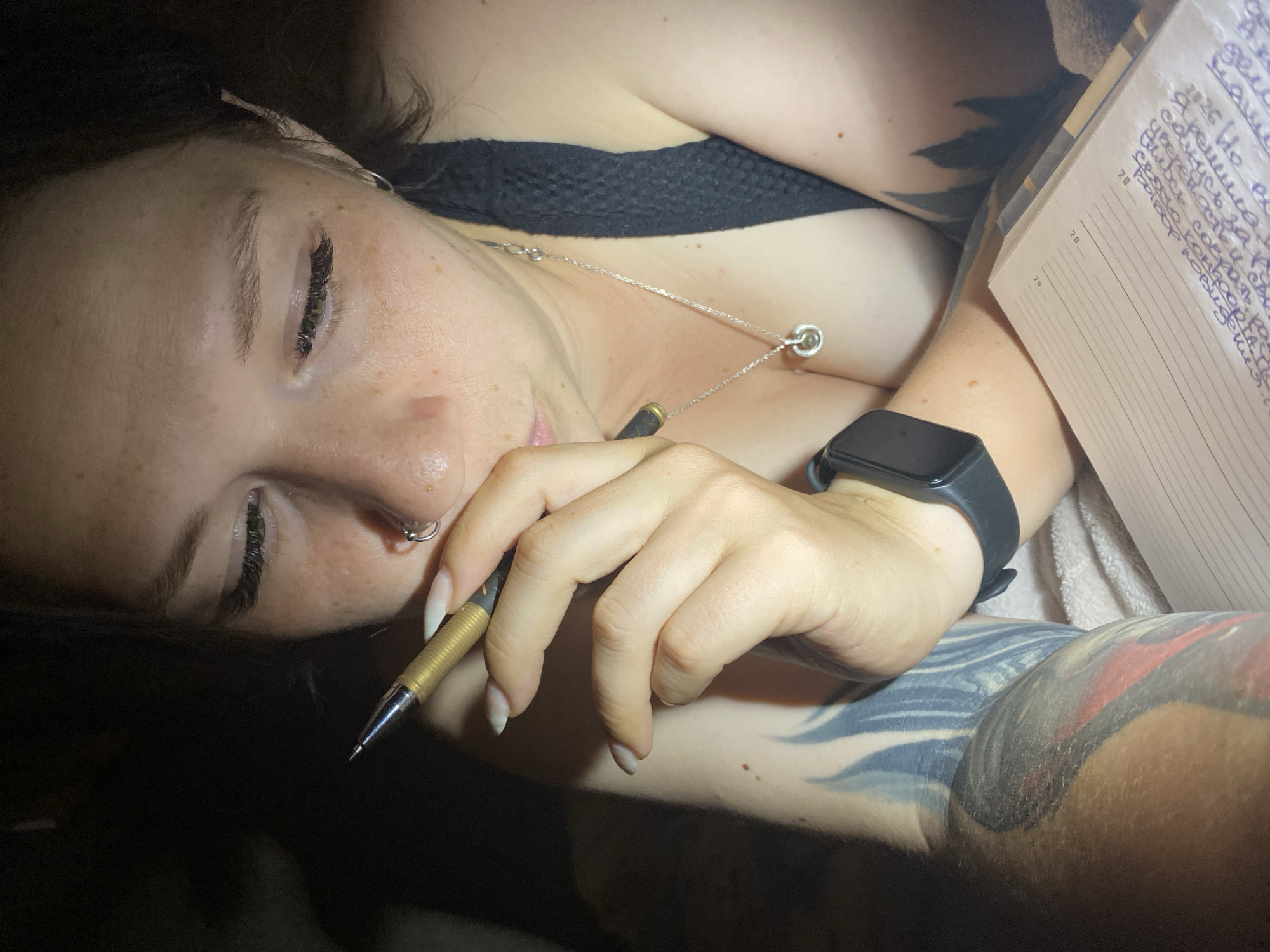 This image is a close-up shot of a woman holding a pen to her lips, with her eyes closed and her head tilted down. She is wearing a black top and a silver necklace with a circular pendant. She has long fingernails, a nose ring, and tattoos on her arm. A notebook and a smartwatch are also visible in the frame. The lighting suggests an indoor setting, possibly at night. The image is set in the city of Chaikovsky, Russia.