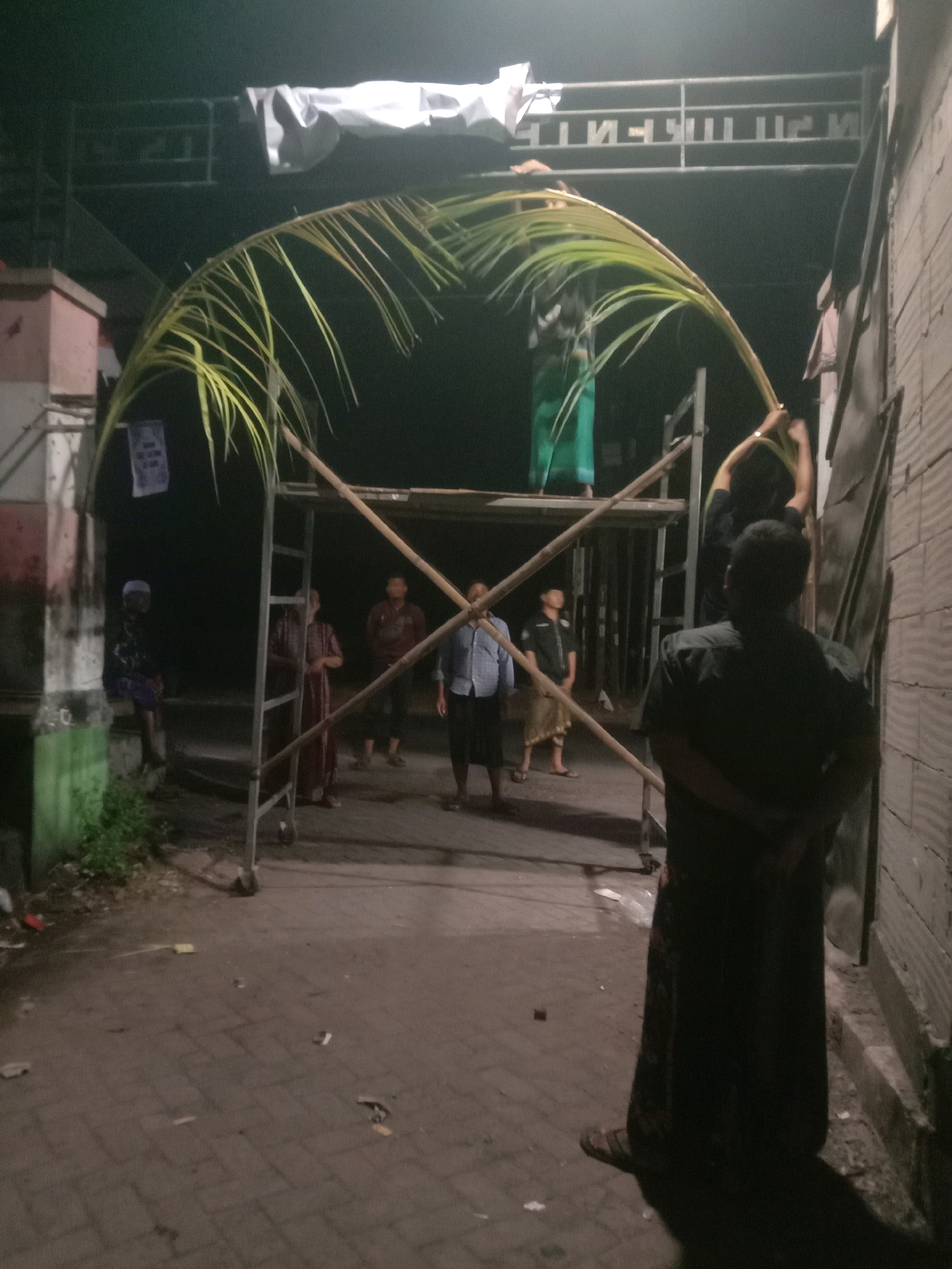 The image depicts an outdoor scene at night, likely in the city of Karang Anyar, Indonesia. A group of individuals are gathered around a scaffolding structure where a large palm frond is being manipulated to form an arch. The arch appears to be part of a temporary decoration or entryway. Several people are visible, some standing on the scaffolding and others on the ground. The ground is paved with interlocking bricks and shows some scattered debris. In the background, a building or structure with a sign is visible, partially illuminated. The overall lighting suggests it is dark, with artificial light sources illuminating the immediate area.