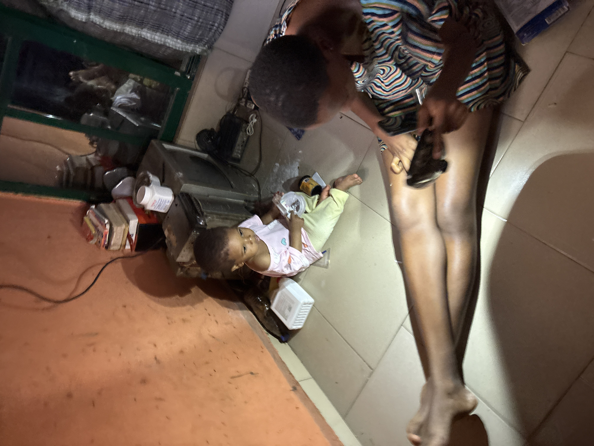 In a dimly lit room, possibly in a residential building in Onitsha, Nigeria, a young child sits on the tiled floor, wearing a light pink and white t-shirt and light yellow pants. The child is looking towards the right side of the frame, with a small, white plastic container and a yellow and brown cylindrical object near them. Adjacent to the child, an adult, partially visible and wearing a colorful striped garment, is seated and appears to be engaged in an activity involving a dark-colored object held in their hands, which rests on their lap. The floor is covered in light-colored square tiles. To the left, a textured, orange-colored wall is visible, with a black electrical cord hanging down. In the background, a window with green framing reveals reflections of interior objects, including what appears to be furniture and possibly a person. Stacked books and a white pill bottle are present on a surface behind the child. The overall impression is of a domestic scene, possibly captured during an evening or in a space with limited natural light.