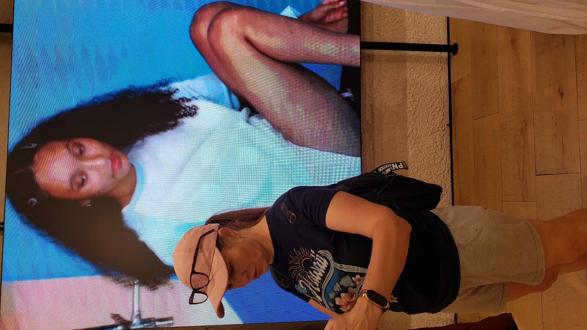 A woman wearing a pink corduroy cap, glasses, a dark t-shirt with a "Hawaii" graphic, and light grey shorts stands in front of a large digital screen displaying a young woman. The screen shows the upper body and legs of the young woman, who has dark, curly hair and is wearing a light blue t-shirt and dark pantyhose. The woman in the foreground is looking down at her phone and wearing a smartwatch on her left wrist. A black backpack is slung over her right shoulder. The background of the screen transitions from a pink and blue abstract pattern to a light blue backdrop. The floor in front of the screen is made of light brown wooden planks. The scene appears to be an indoor retail or exhibition space, possibly in the city of Prešov, Slovakia.