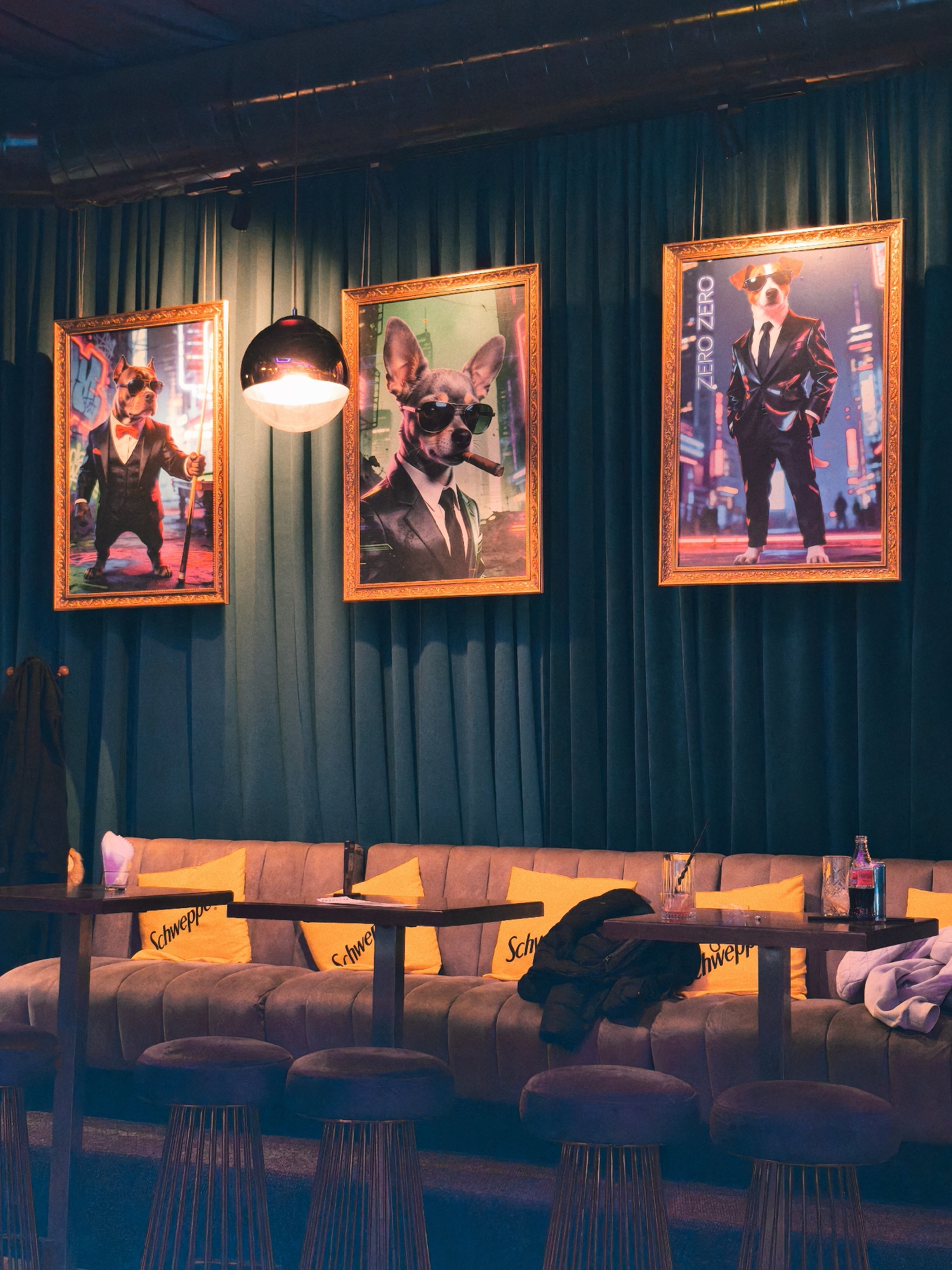 Indoor scene in a bar setting with three framed dog portraits hanging on a dark teal draped wall. The frames are gold colored. The portraits depict anthropomorphic dogs wearing sunglasses and dressed in suits. The portrait on the left shows a dog with a bow tie holding a pool cue. The center portrait shows a dog with a cigar. The portrait on the right has the words "ZERO ZERO ZERO" above the dog. Below the portraits is a long, gray tufted couch with yellow throw pillows that say "Schweppe". Several cocktail tables with wire-framed stools in front of the couch. A half-empty glass, a bottle of Coca-Cola, a black jacket and a white sweatshirt or t-shirt are on the table or couch. A coat hangs on a coat rack on the left side of the image. A modern light fixture hangs between the first and second portrait. The lighting is dim with a blue tint. The specific location in Varna, Bulgaria is not visible in the image.
