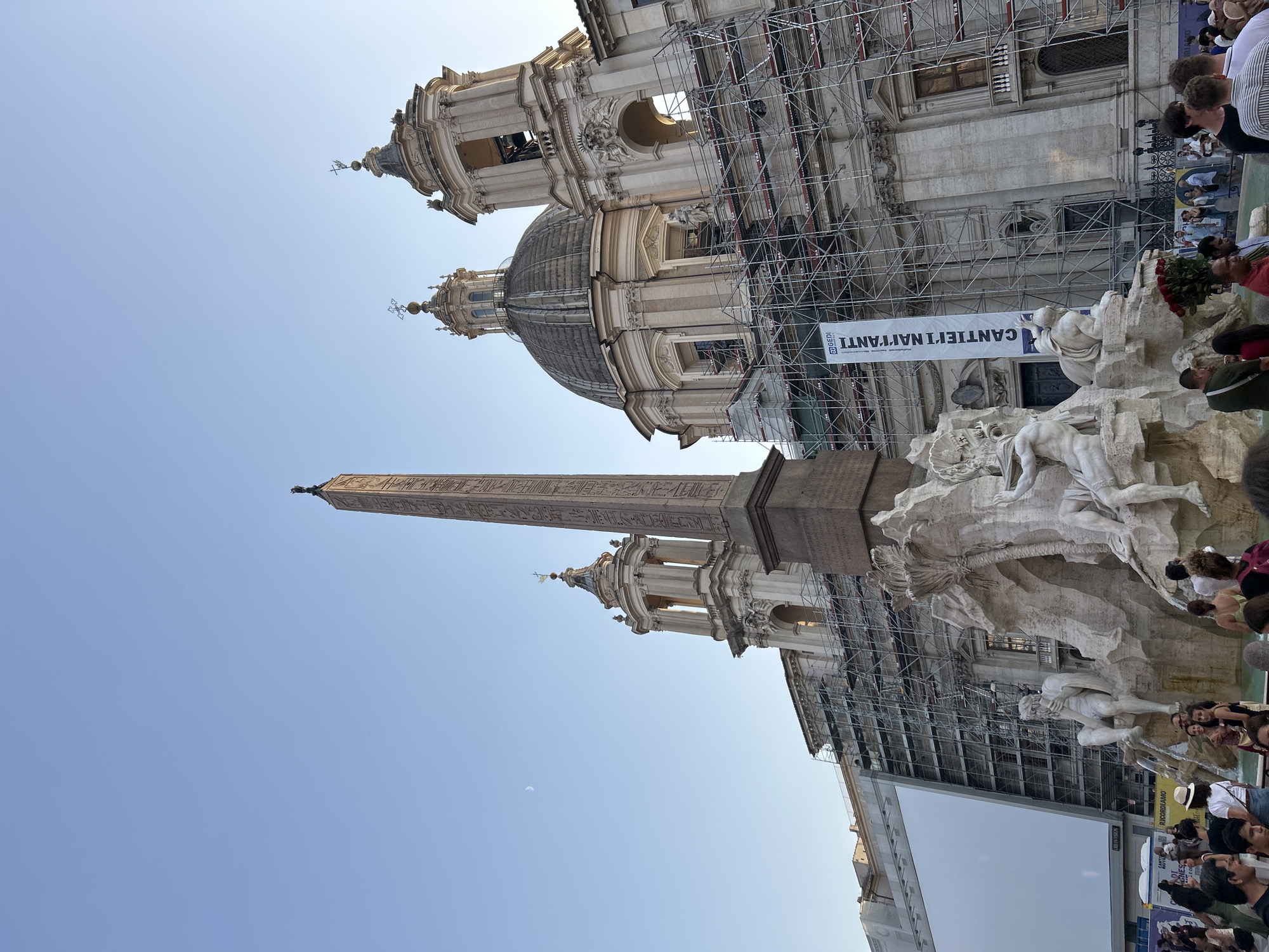 This is a photograph taken from a low angle, looking up at an obelisk and a church facade covered in scaffolding. The sky is clear and blue, suggesting a sunny day. The image appears to be a wide shot of a public square in Rome, Italy, as indicated by the architectural style of the church and the presence of an obelisk, a common feature in Roman public spaces.
The main subjects are the architectural elements, specifically the towering obelisk and the ornate church with its dome and bell towers. The scaffolding is a prominent feature, indicating ongoing construction or renovation work on the church. A large white banner hangs from the scaffolding, with visible text that reads "CANTIERE IN CORSO" which translates to "Construction site in progress" in Italian. Further text is visible on another banner near the bottom of the image.
In the foreground, a large fountain is visible, featuring several white marble sculptures of human figures and mythical creatures. These sculptures appear to be part of a Baroque-style fountain, a characteristic of Roman fountains. A crowd of people are gathered around the fountain, some looking at the sculptures, others taking pictures. Their presence suggests this is a popular tourist attraction.
The time of day appears to be late afternoon or early evening, given the angle of the sunlight casting shadows. The overall mood is one of lively public activity, with tourists and locals coexisting amidst the grand historical architecture and ongoing restoration work. The presence of the moon, a faint crescent, is also visible in the sky, adding to the sense of time and place.