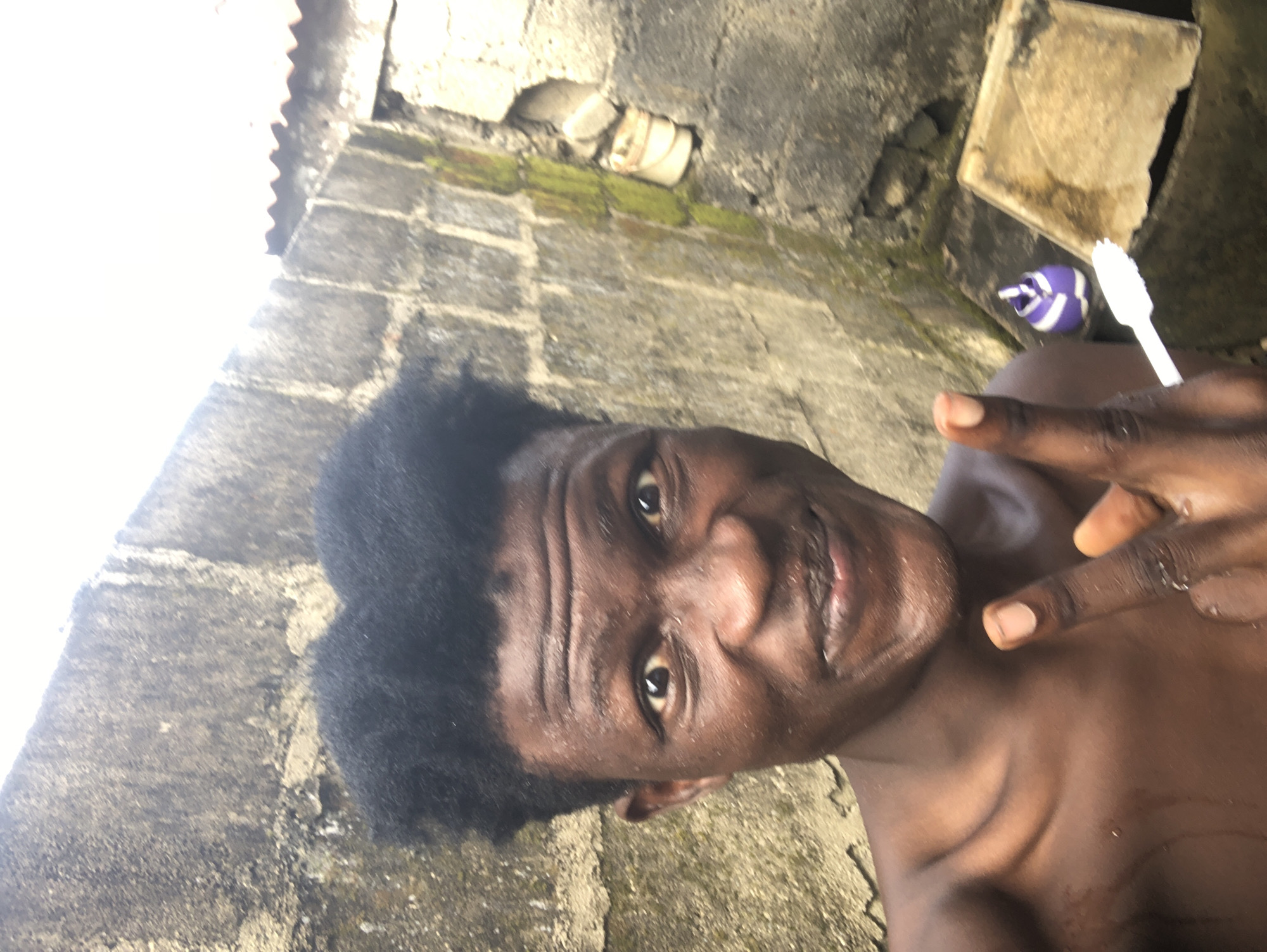 The image is a close-up selfie of a young Black male with dark skin and short, black, curly hair, taken outdoors. He is positioned in the foreground, his head and shoulders filling most of the frame. He has wide, dark eyes, a broad nose, and a wide smile, revealing his teeth. His skin appears wet or dewy. He is wearing dark, possibly wet, clothing, though only his upper chest and shoulders are visible. He is making a peace sign with his right hand and pointing his left index finger upwards.
The background consists of a rough, textured concrete wall with visible signs of weathering, including patches of green moss. To the right of the man, in the mid-ground, there is a concrete structure, possibly a sink or drain, with a small, purple and white object (possibly a small bottle or container) and a white toothbrush lying nearby. A corrugated metal sheet is visible along the top right edge of the frame, suggesting an outdoor setting.
The lighting suggests it is daytime, with a bright, possibly overcast sky. The overall environment appears to be a somewhat rudimentary outdoor space. The man's expression and pose suggest a casual and perhaps playful demeanor. The presence of the toothbrush might indicate that the photograph was taken while he was in the process of or had just finished performing personal hygiene. The environment, particularly the unfinished concrete and outdoor plumbing fixture, could be indicative of a residential area in Ado, Nigeria, where such construction and sanitation setups are common in many parts of the city.
