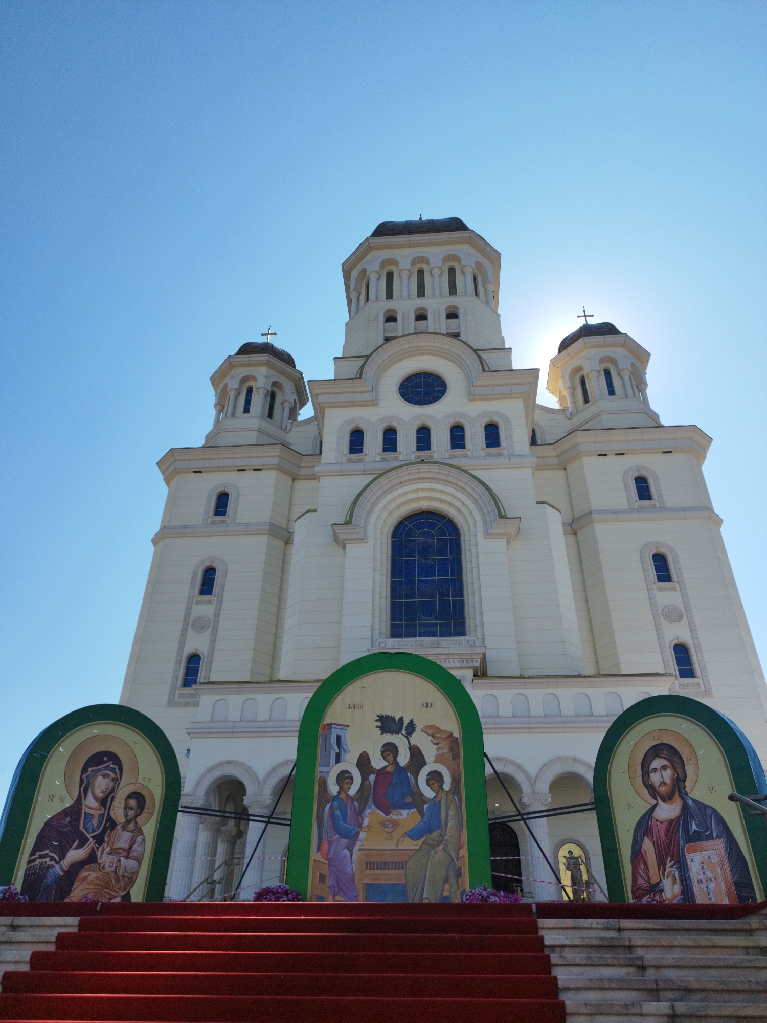 The photograph captures the exterior of the Cathedral of the Salvation of the Nation in Bucharest, Romania. The imposing cream-colored building features multiple towers with dark domes topped with crosses, and numerous arched windows. In the foreground, a red carpet leads up a wide set of stairs. Flanking the stairs are three large, arched religious icons set against green backdrops. The icon on the left depicts the Virgin Mary holding the Christ Child, the central icon shows a biblical scene with three figures, and the icon on the right features Jesus Christ holding a book. The sky above is clear and blue, with the sun shining brightly and casting a lens flare near the top of the central tower.