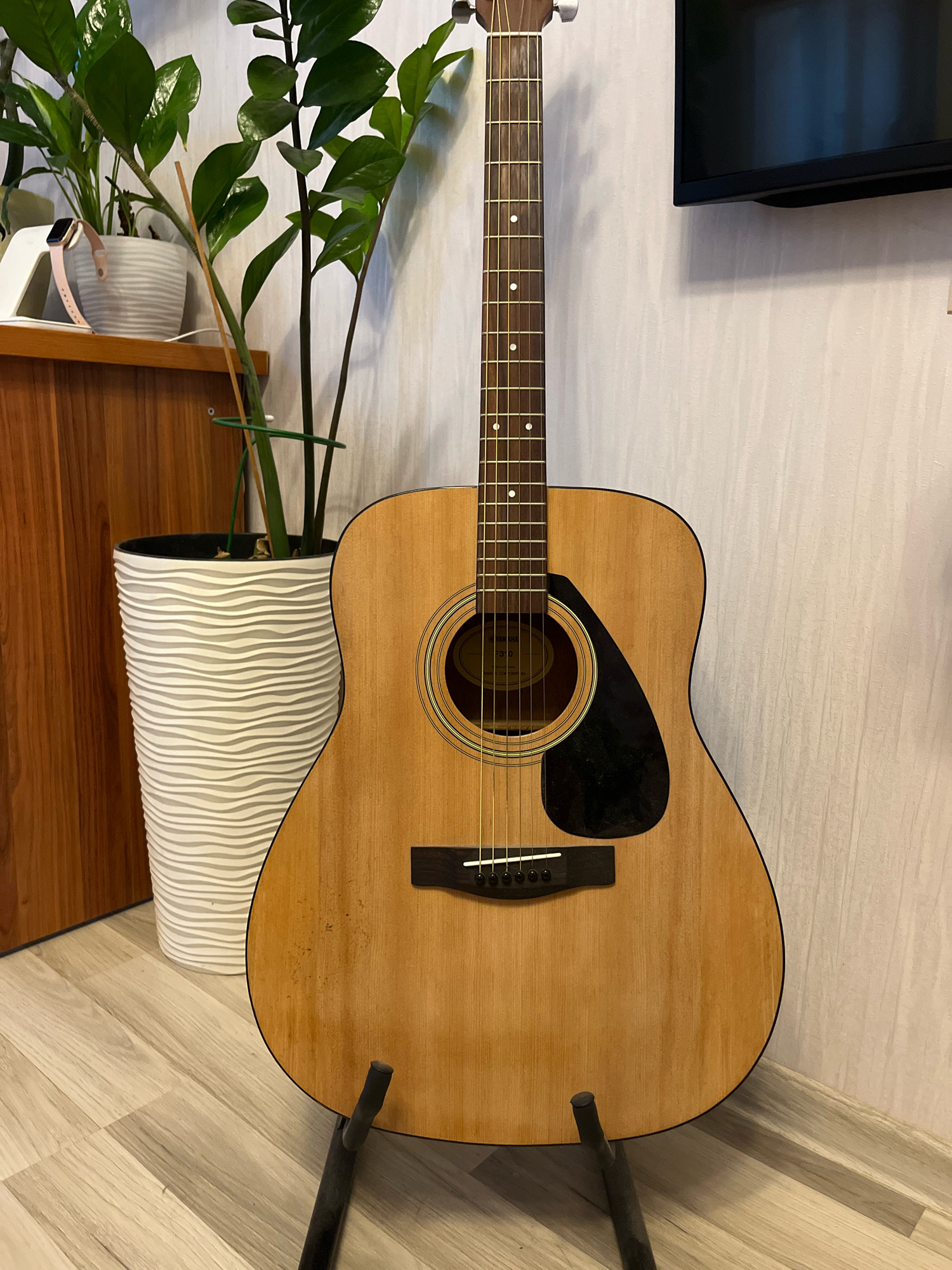 An acoustic guitar is positioned on a black stand in the center of the frame, with its body facing the viewer. The guitar has a light wood finish, a black pickguard, and six strings. To the left of the guitar, there is a large white textured planter with green plants. Behind the guitar, a textured wall is visible, with a portion of a television screen in the upper right corner. The floor is made of light-colored wooden planks. The scene appears to be indoors, possibly a living room. The city of Chaikovsky, Russia, is not depicted or referenced in the image.